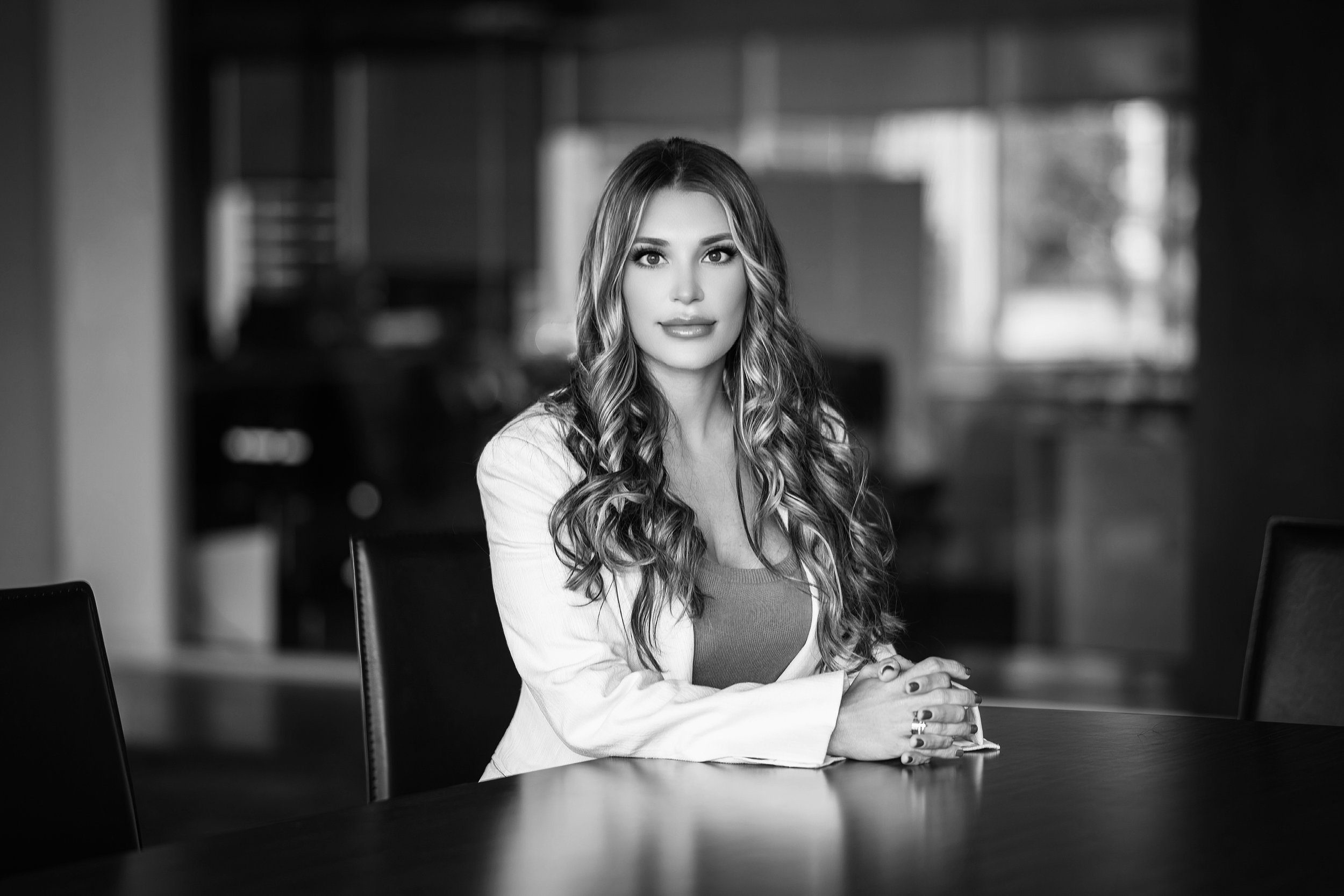 A woman with long wavy hair, wearing a light blazer over a top, sitting at a table in a modern office or meeting room, looking directly at the camera.