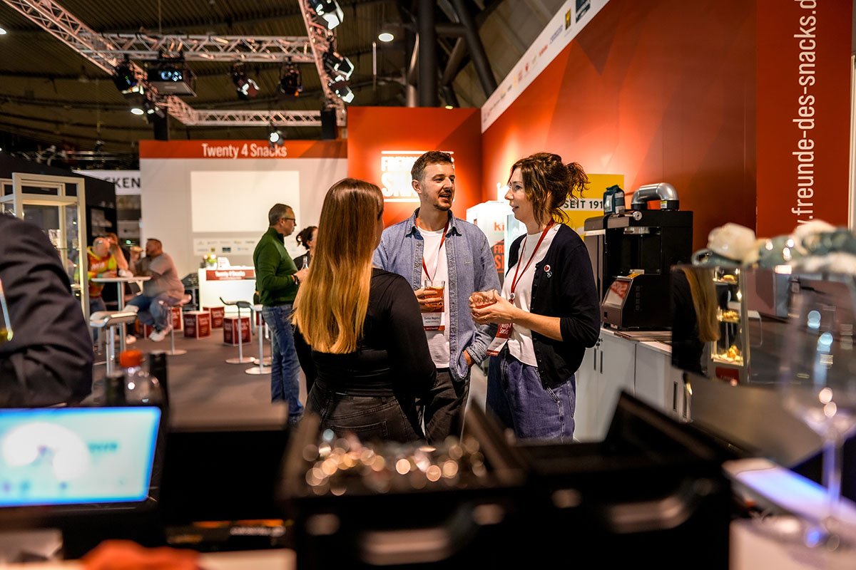 Drei Personen unterhalten sich an einem Messestand mit der Aufschrift 'Twenty 4 Snacks' bei einer Messe. Im Hintergrund sind weitere Besucher und Aussteller sichtbar.