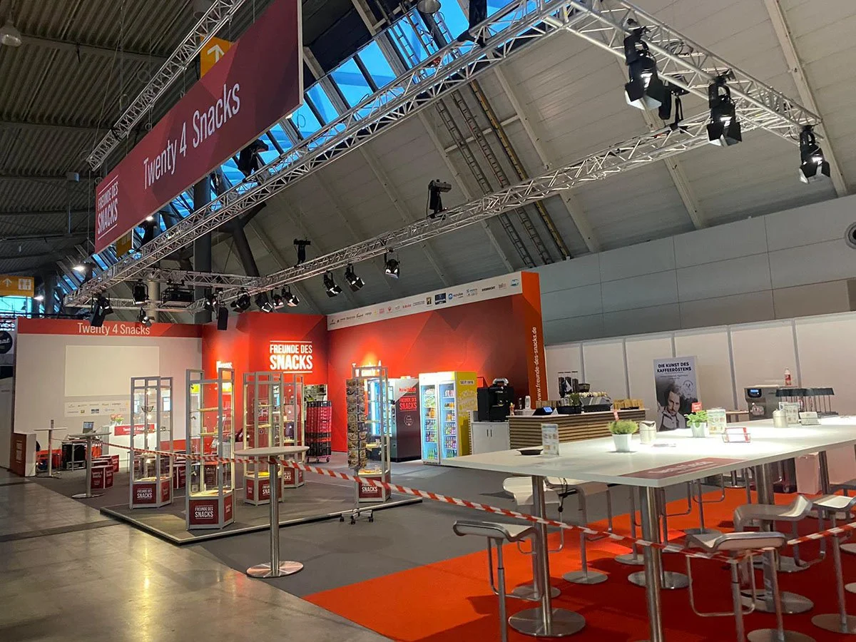 Verkaufsstand mit Snacks und Getränke in einer Konferenzhalle, mit Tischen und Sitzgelegenheiten.