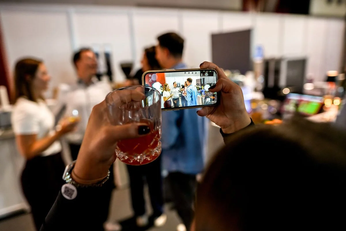 Person fotografiert eine Gruppe von Menschen mit einem Smartphone bei einer Veranstaltung, während sie ein Glas mit rotem Getränk in der Hand hält.