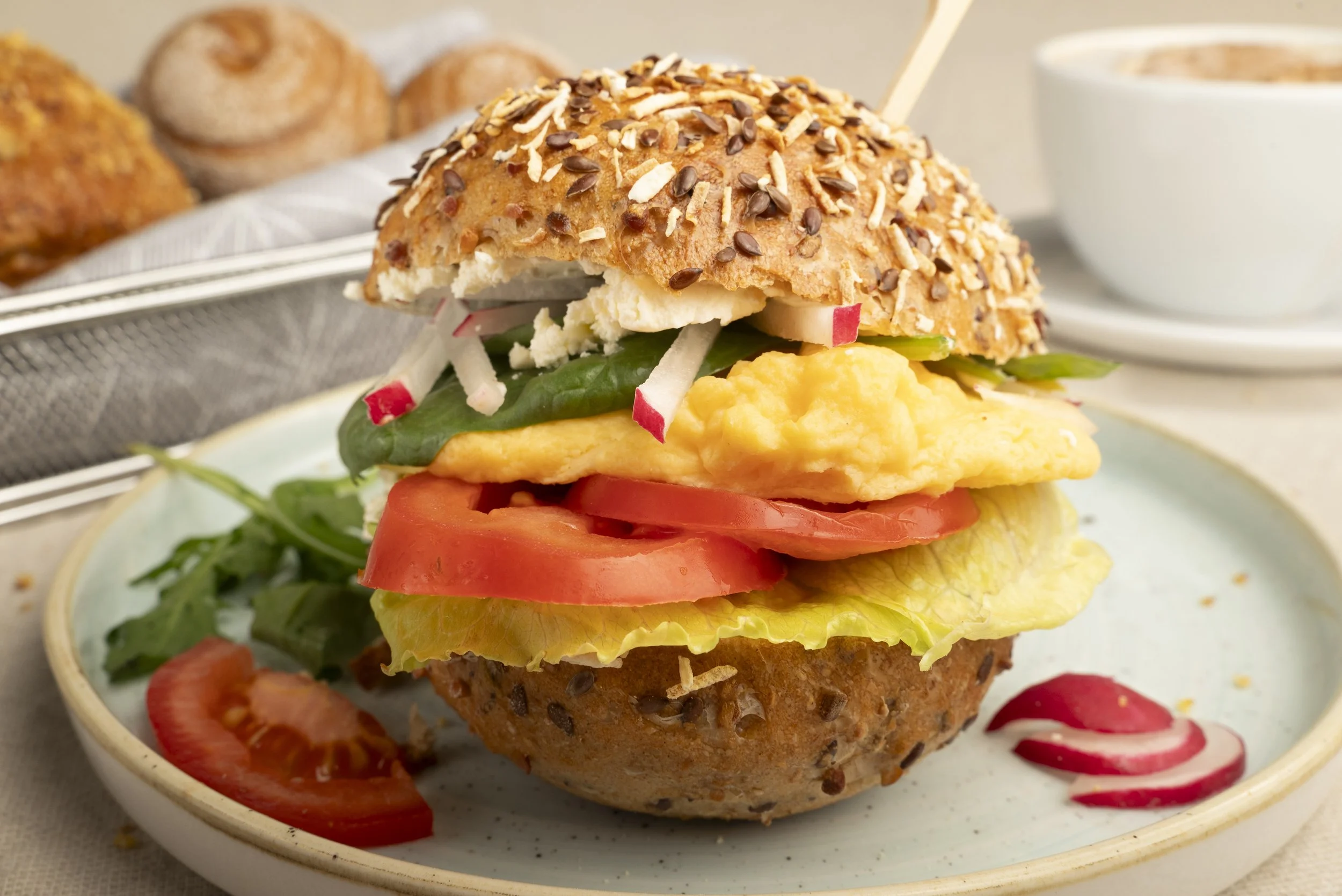 Vielstöckiger glutenfreier Sandwich mit Ei, Tomaten, Salat, Radieschen, auf einem Teller, mit Marmelade und Brötchen im Hintergrund.