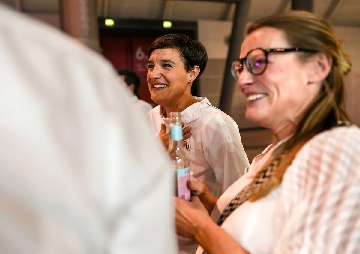 Zwei lachende Frauen im Gespräch bei einer Veranstaltung, eine hält ein Getränk in der Hand.