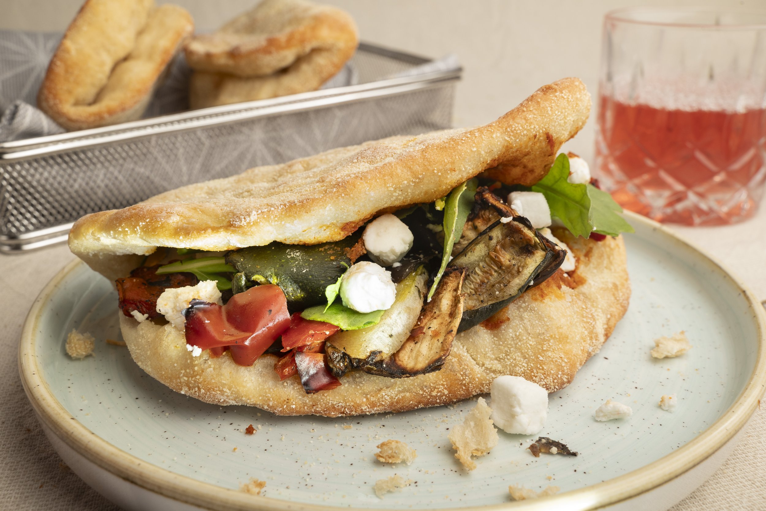 Fischsandwich mit Gemüse auf einem Teller, im Hintergrund ein Glas Rosé-Wein und ein Korb mit frittiertem Teiggebäck