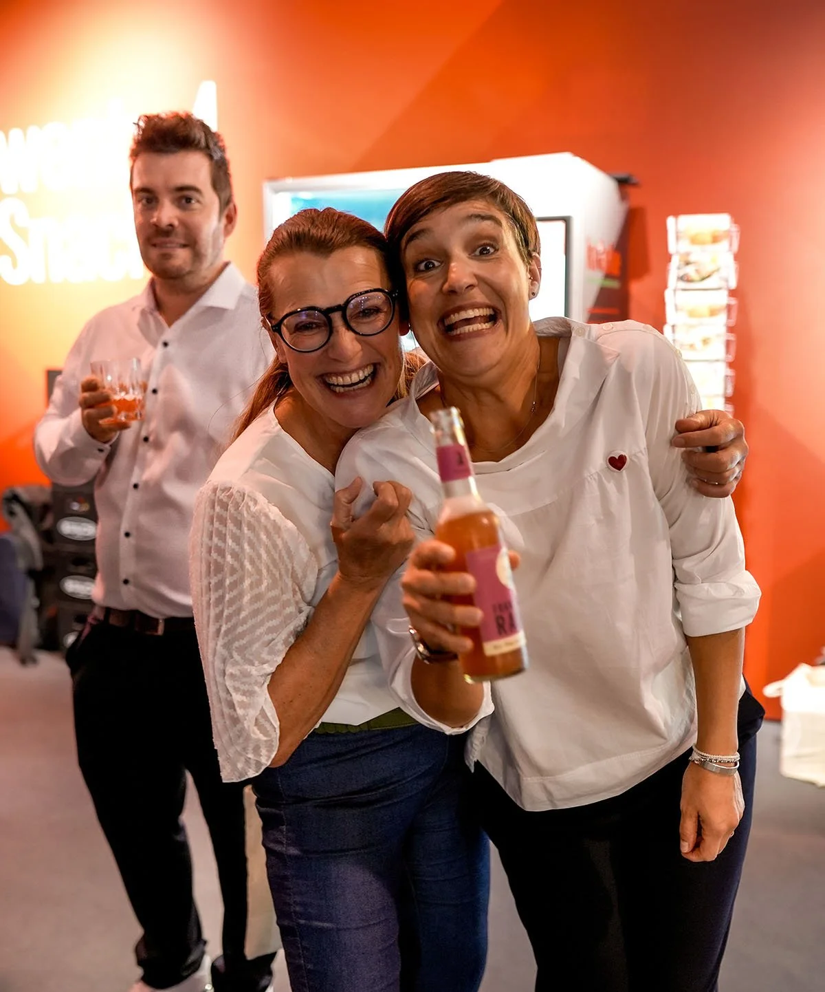 Drei Menschen lachen und stoßen mit Getränken an, im Hintergrund eine Person ebenfalls mit Getränk, in einer fröhlichen Atmosphäre.