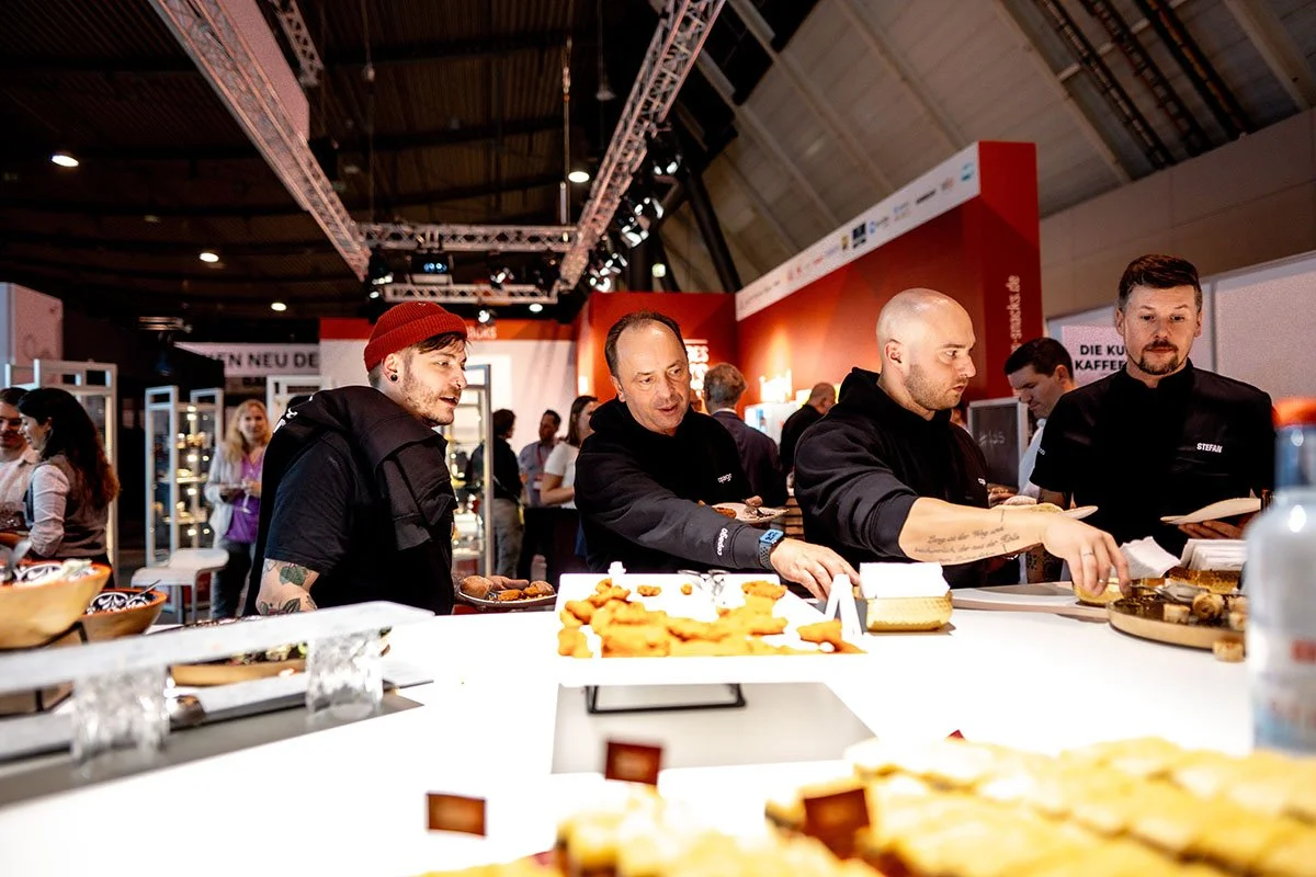 Menschen am gastronomischen Stand auf einer Messe prüfen Speisen in einem Innenraum mit roten Wänden und hohen Decken.