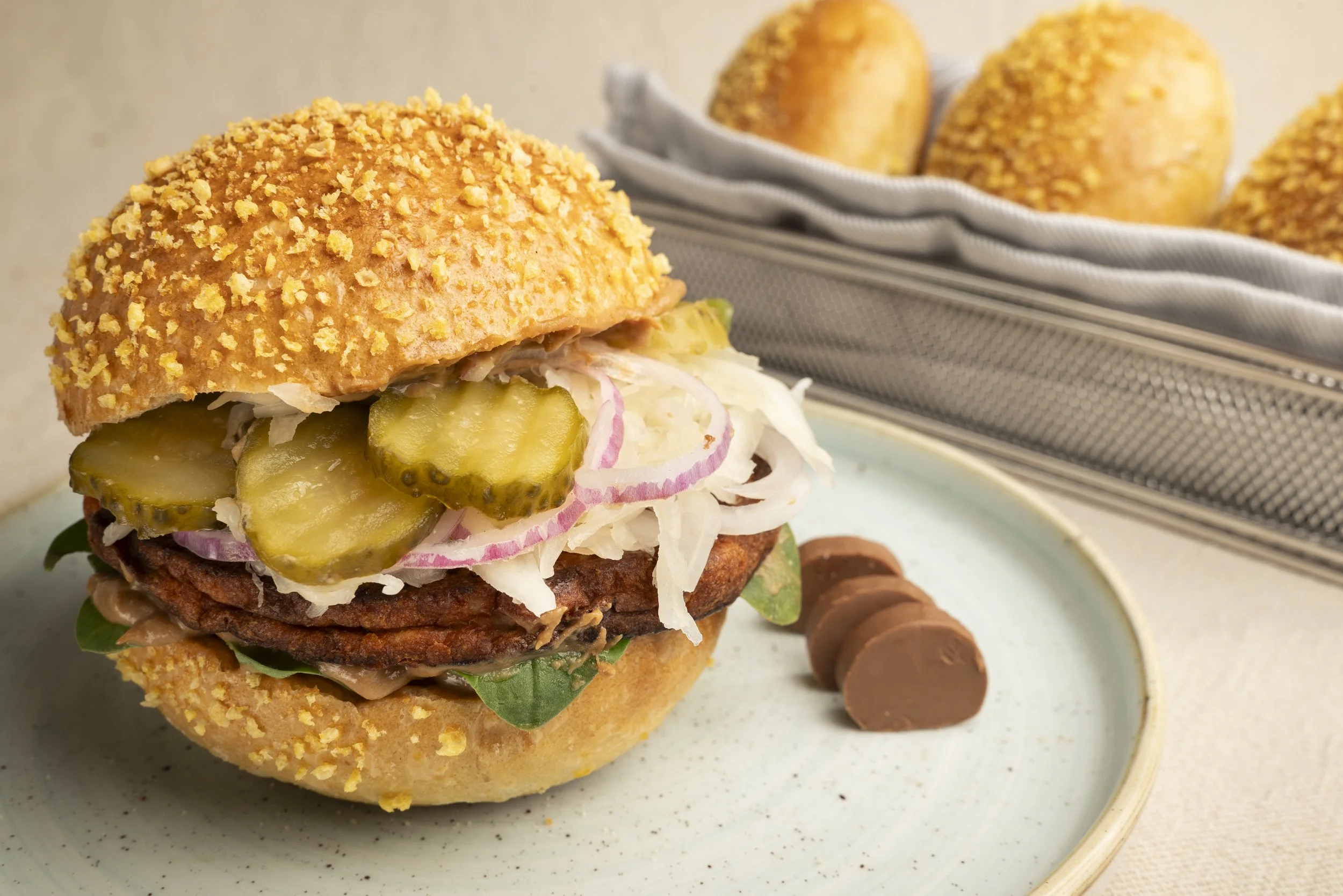 Ein Hamburger mit Gurken, Zwiebeln, Kohl und einem Sesambrötchen auf einem weißen Teller, daneben drei Schokoladenscheiben, im Hintergrund ein Korb mit weiteren Brötchen.