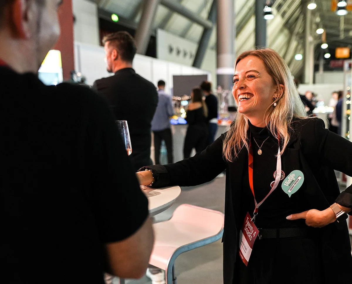 Eine lachende Frau mit lila Haare, die auf einer Messe mit einem Namensschild spricht, während im Hintergrund andere Messebesucher sind.