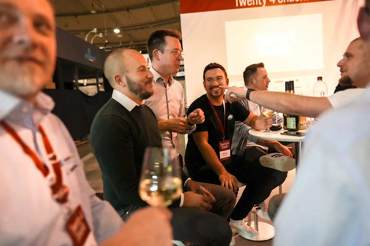 Gruppe von Männern, die bei einer Veranstaltung lachen und Drinks trinken, im Hintergrund ein Bildschirm mit einem Präsentationsbild.