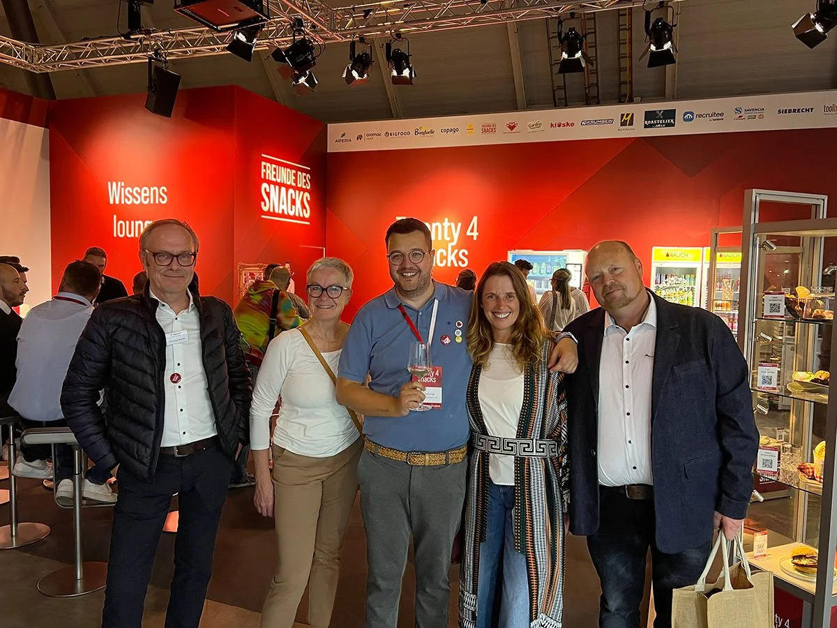 Gruppe von fünf Menschen, die bei einer Veranstaltung stehen, mit einem roten Hintergrund, auf dem steht 'FREUNDE DES SNACKS' und weitere Texte. Einige Personen lächeln in die Kamera, eine Person hält ein Weinglas.
