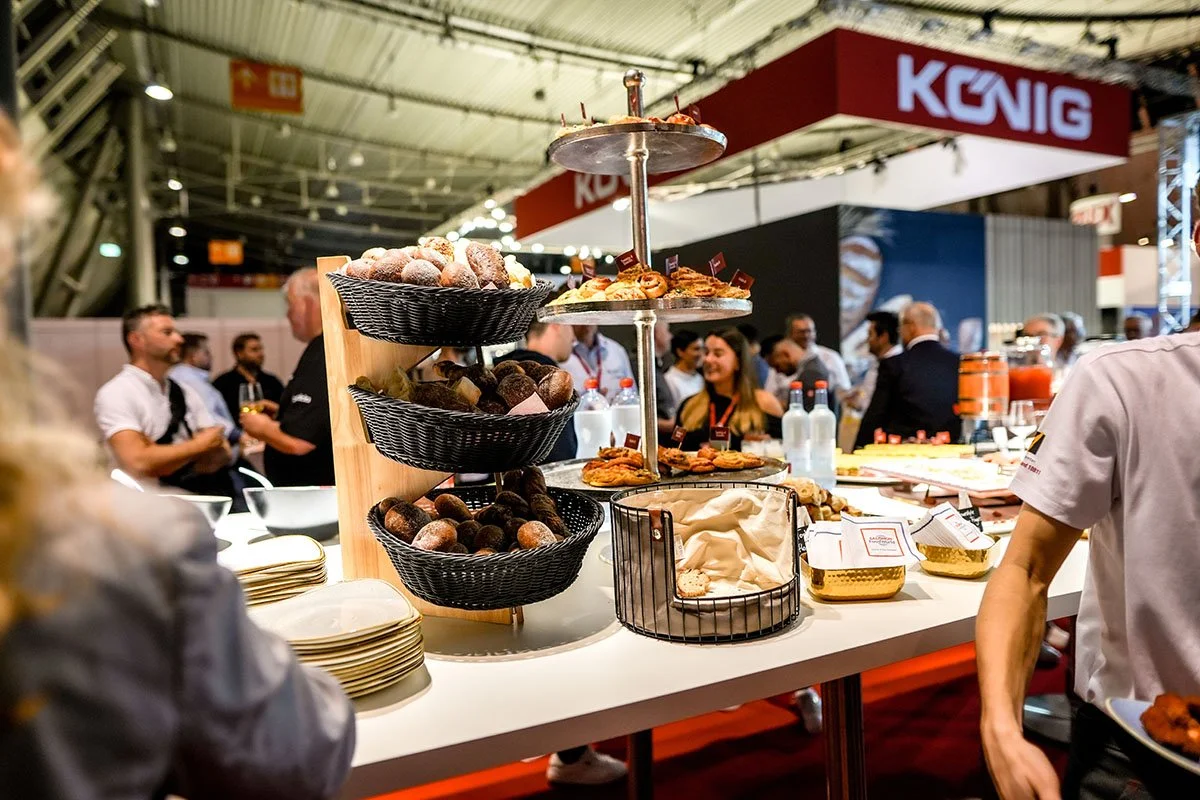 Buffet mit Süßigkeiten und Gebäck bei einer Messe, im Hintergrund Menschen im Gespräch und ein großes »KÖNIG«-Schild.