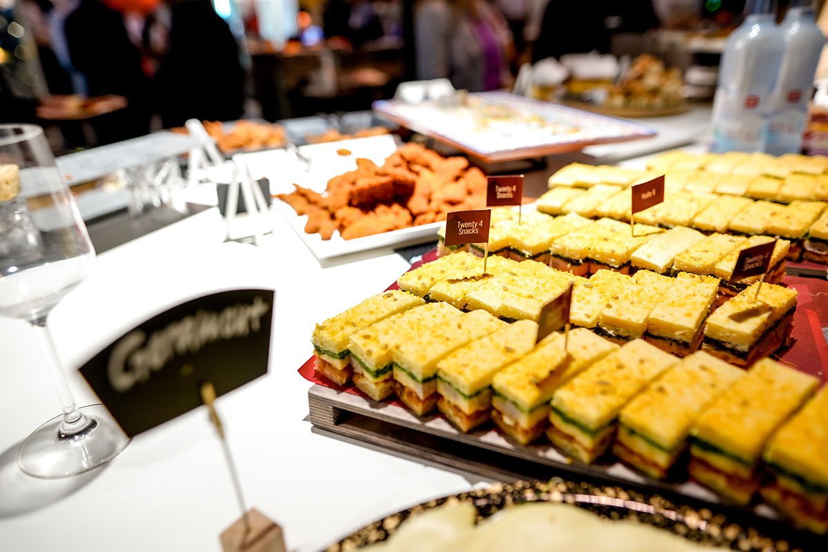 Verschiedene belegte Sandwiches und Snacks auf einem Buffet-Teller, im Hintergrund weitere Snacks und Getränke, bei einer Feier oder Veranstaltung.