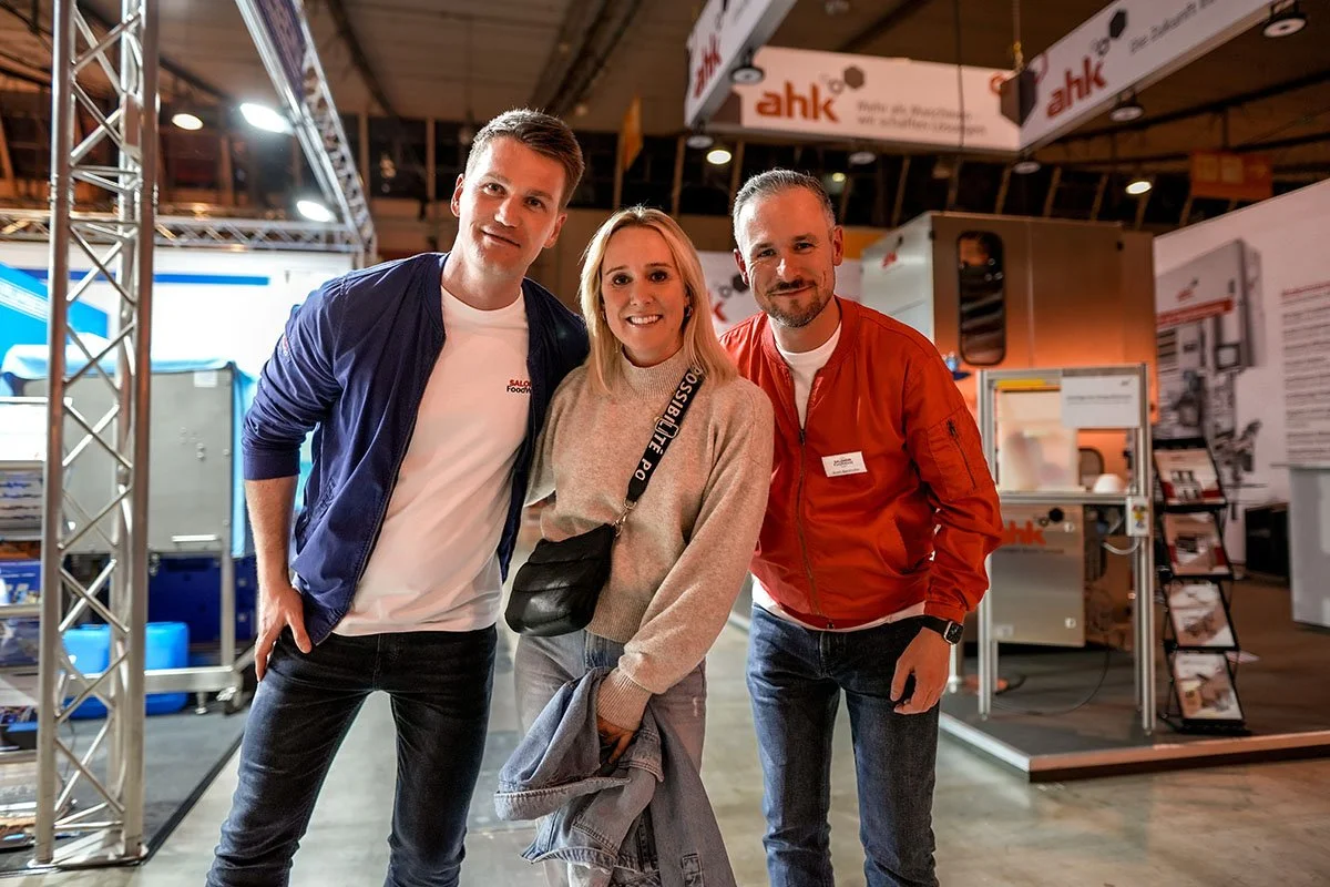 Drei Personen, zwei Männer und eine Frau, posieren auf einer Messe vor Messeständen. Alle lächeln. Der Mann auf der linken trägt eine blaue Jacke und ein weißes T-Shirt, die Frau in der Mitte trägt einen grauen Pullover, und der Mann auf der rechten 