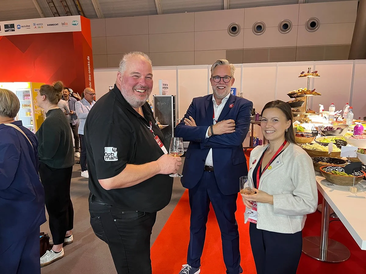 Drei Menschen mit Getränken im Gespräch auf einer Messe oder Veranstaltung, im Hintergrund Essensbuffet mit Salaten und Snacks, im Raum Hohlraumlüftung und verkleidung an der Decke.