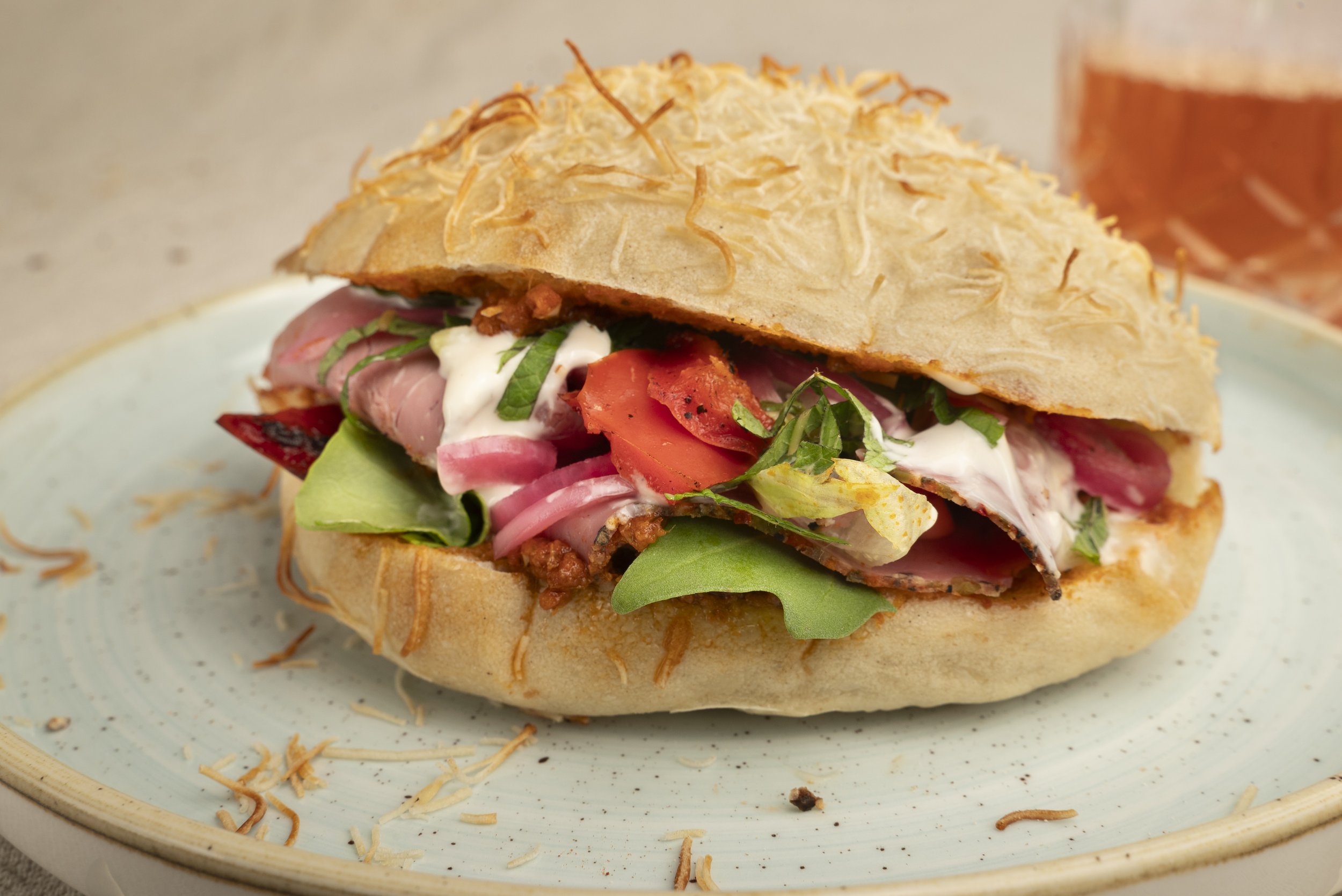 Ein belegtes Ciabatta-Brötchen mit frischen Kräutern, Salat, Tomaten, Zwiebeln, veganem Hackfleisch, und Käse, serviert auf einem weißen Teller.