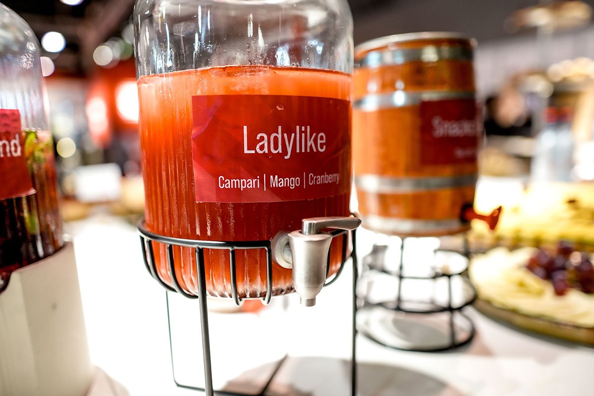 Ein Glaskrug mit rotem Getränk, gekennzeichnet als LadyLke mit Geschmackssorten Campari, Mango und Cranberry, stehend auf einem Metallgestell.