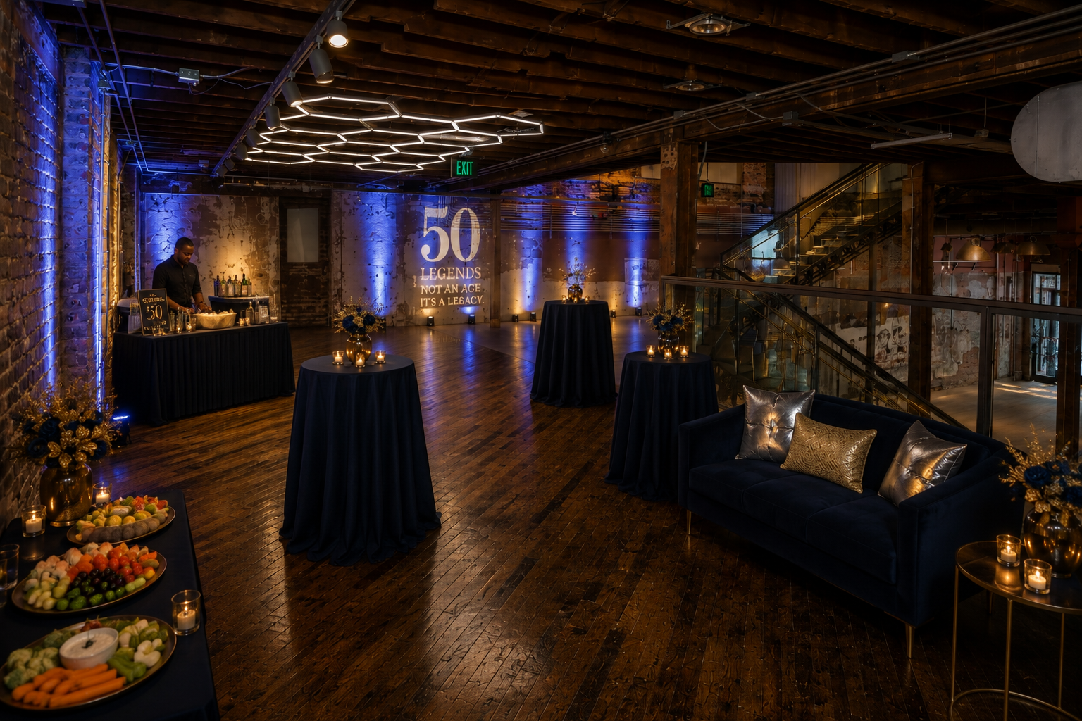 Elegant event space decorated with dark blue tablecloths, floral arrangements, and candles, celebrating 50 years with the message '50 Legends, Not an Age, It's a Legacy' projected on the wall. There are high-top tables, a sitting area with a dark blue sofa and pillows, a bar area, and a dessert table with fruit and snacks. The space has wooden floors, exposed brick walls, and modern lighting.