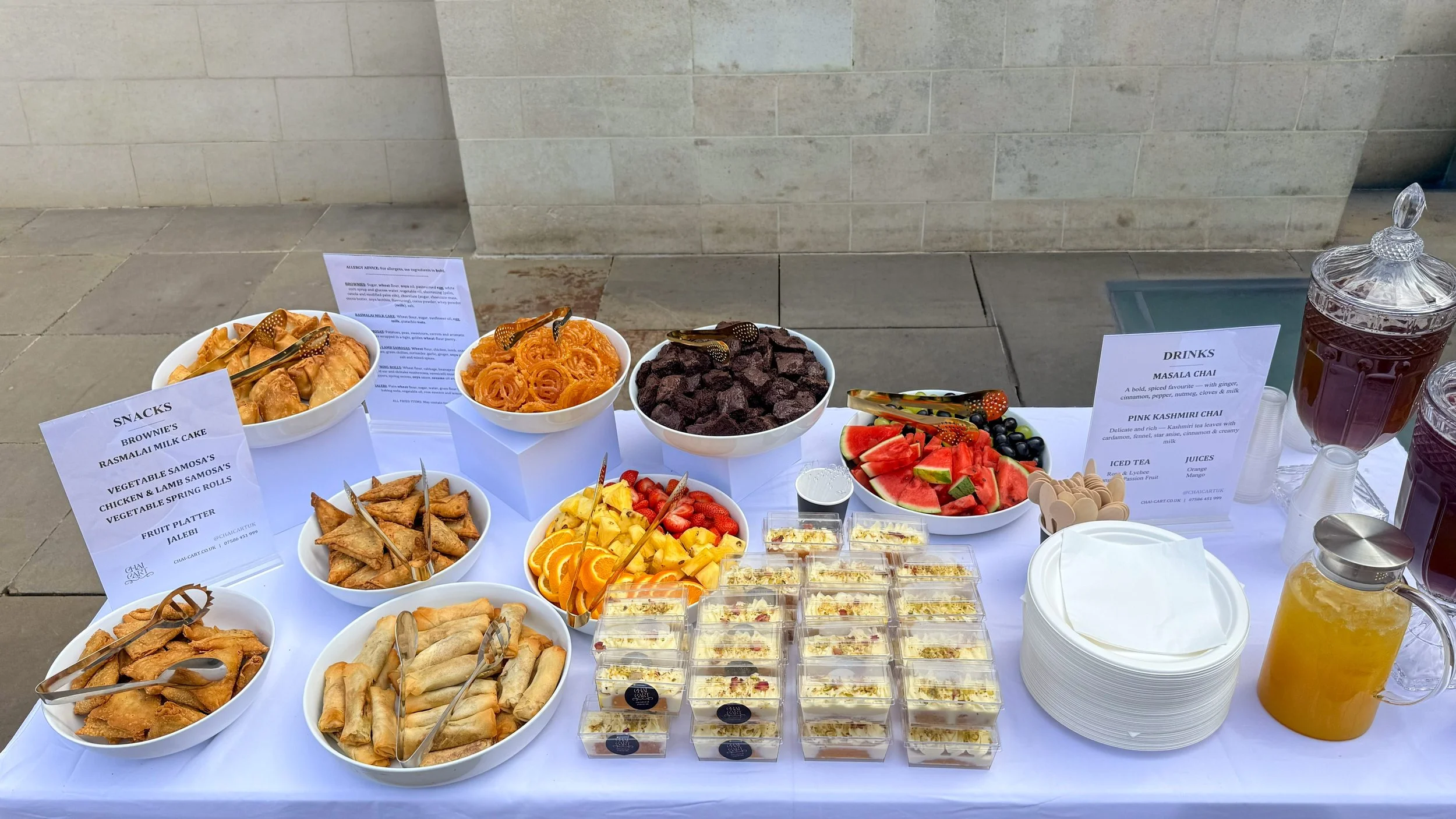 A buffet table with bowls of samosas, fruit, and chocolate-covered treats, along with juice and water, and a sign indicating assorted snacks and drinks.