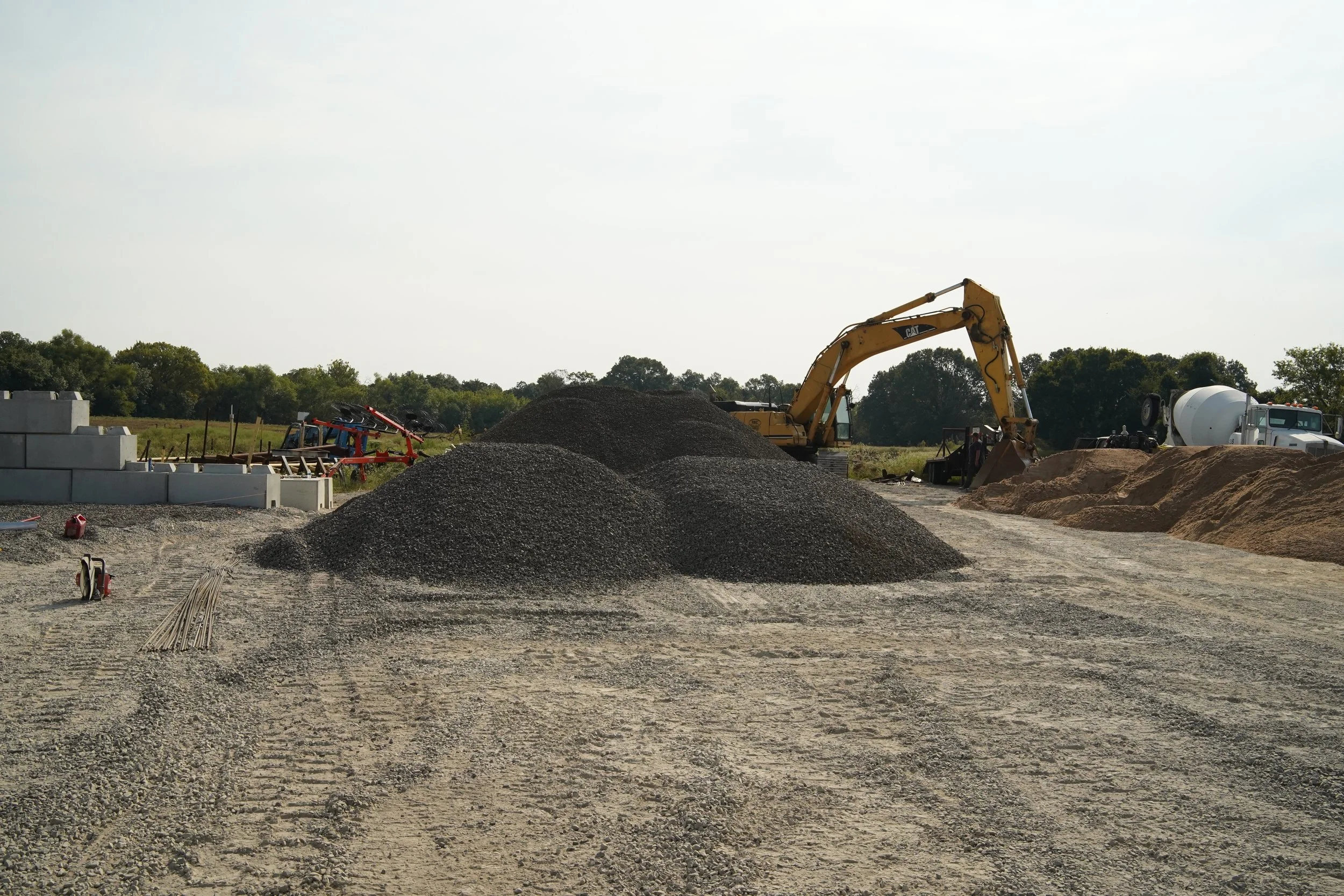Construction site with piles of gravel and sand, an excavator, a cement mixer truck, and construction materials under a cloudy sky.