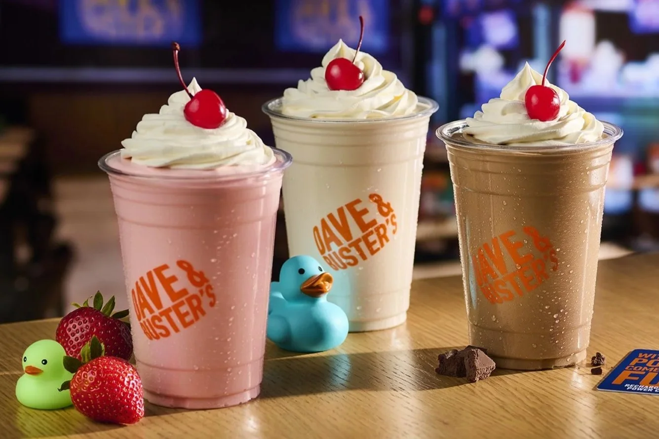 Three shakes topped with whipped cream and cherries on a wooden table, with strawberries, rubber duckies, and chocolate pieces nearby, inside a Dave & Buster's restaurant.