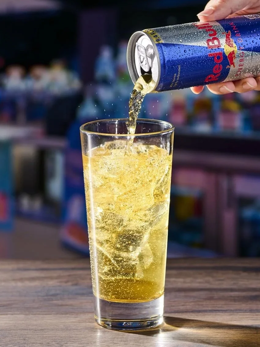 A hand pouring Red Bull energy drink into a tall glass with ice on a wooden surface, with a blurred background of a store.