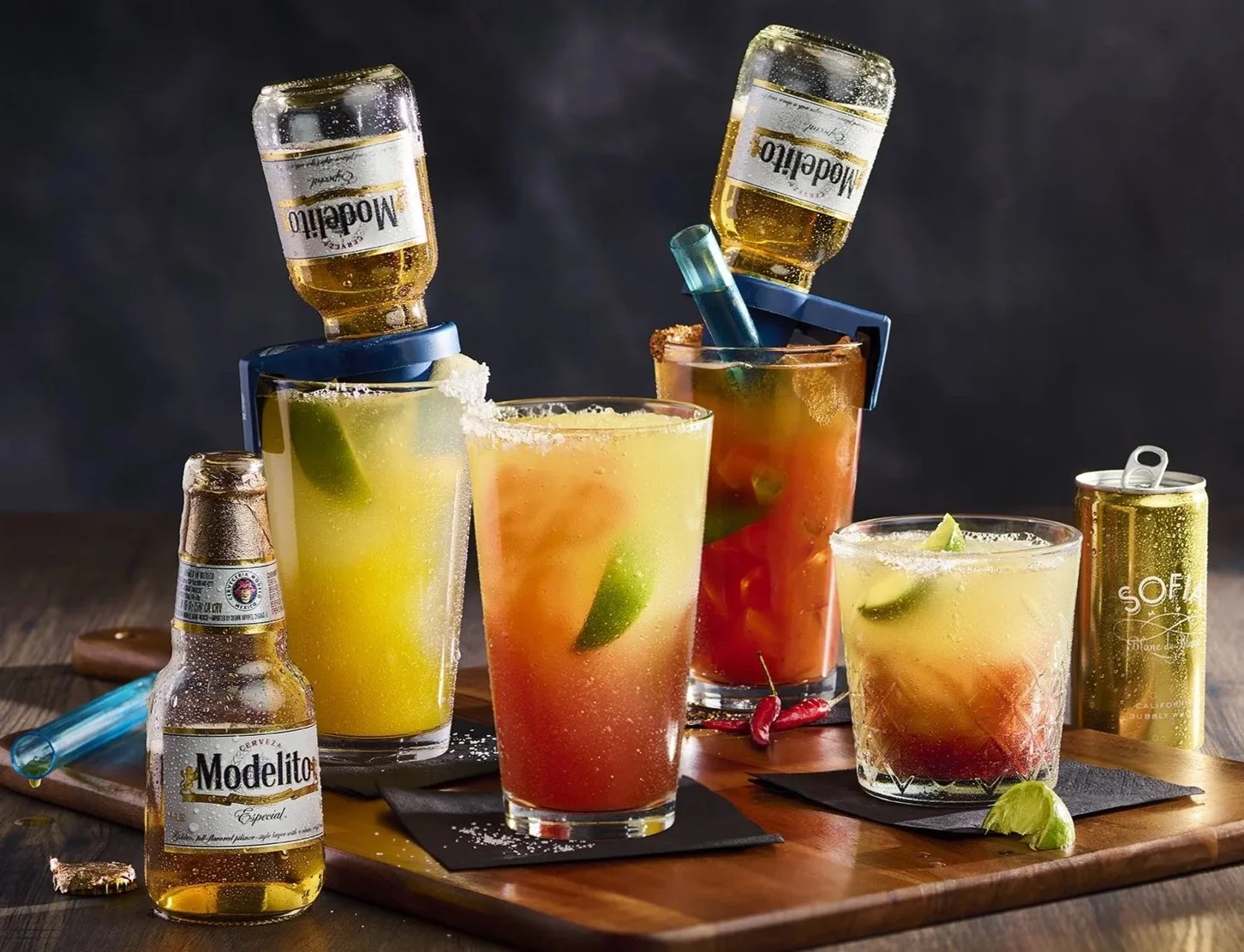 Assorted cocktails with lime wedges and chili peppers on a wooden board, some with beer bottles and cans, garnished with straws and salt rims.