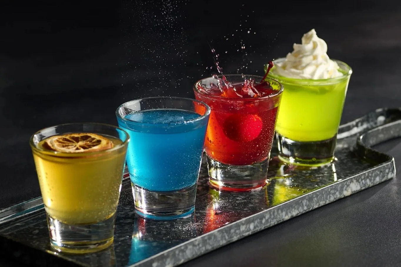 Four colorful cocktails with garnishes on a reflective metal tray against a black background.
