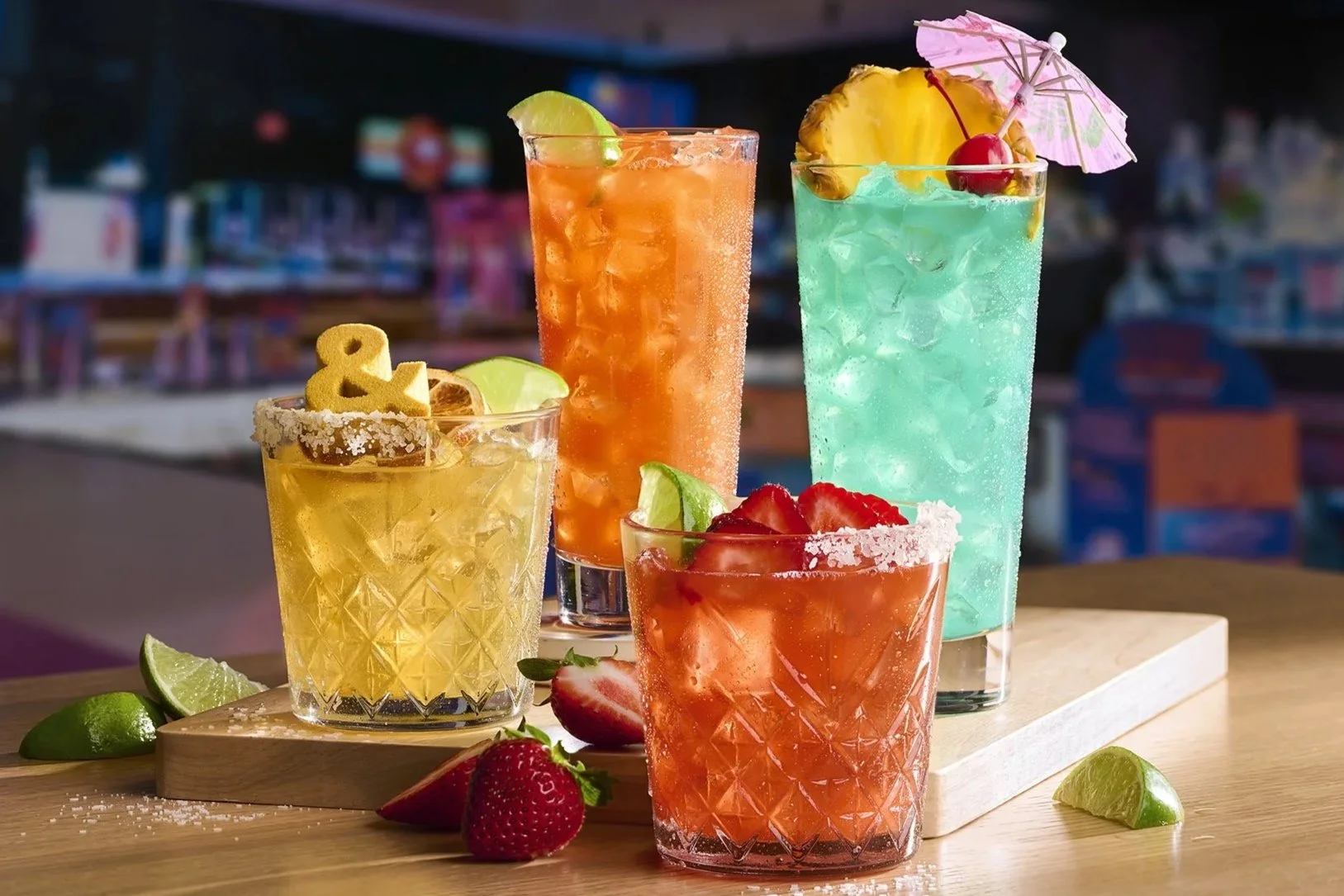 Four colorful cocktails with fruit garnishes on a wooden serving board, with strawberries, lime wedges, and salt on the table, in a bar or outdoor setting.