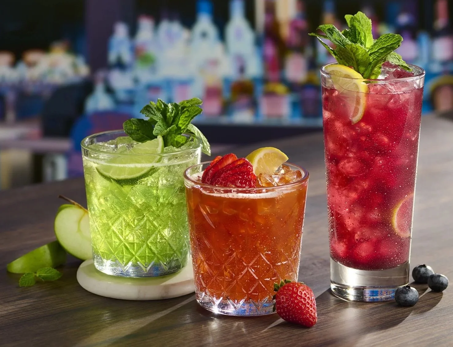 Three colorful cocktails on a wooden table with berries and fruits around, with a blurred bar background.