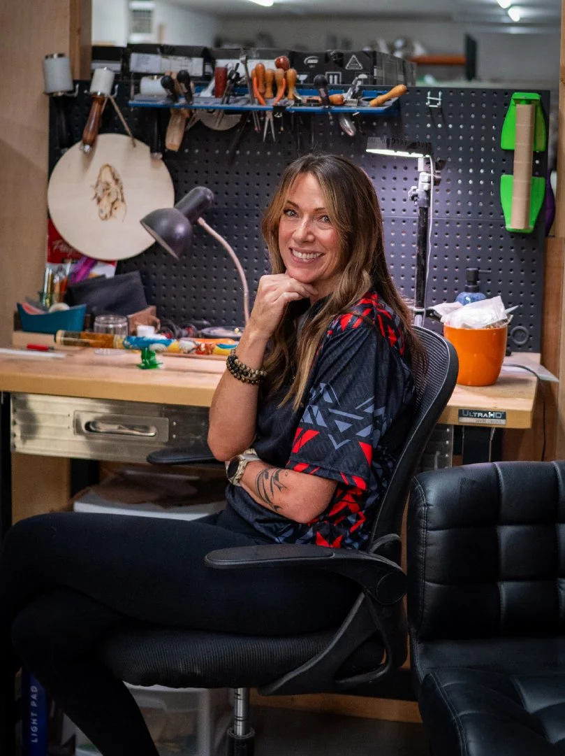 A woman with long brown hair smiling and sitting on a black office chair in a workshop. Behind her is a pegboard with tools hanging, including screwdrivers, pliers, and a saw, with a workbench cluttered with various items. The workshop has focused lighting and a casual, creative atmosphere.