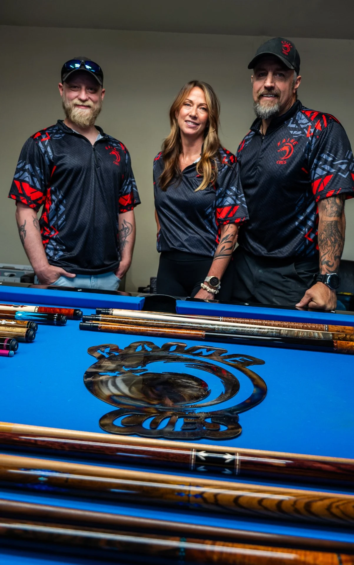 Three people standing behind a pool table with pool cues and a decorative logo on the table. They are wearing matching black sports jerseys with red and gray patterns.