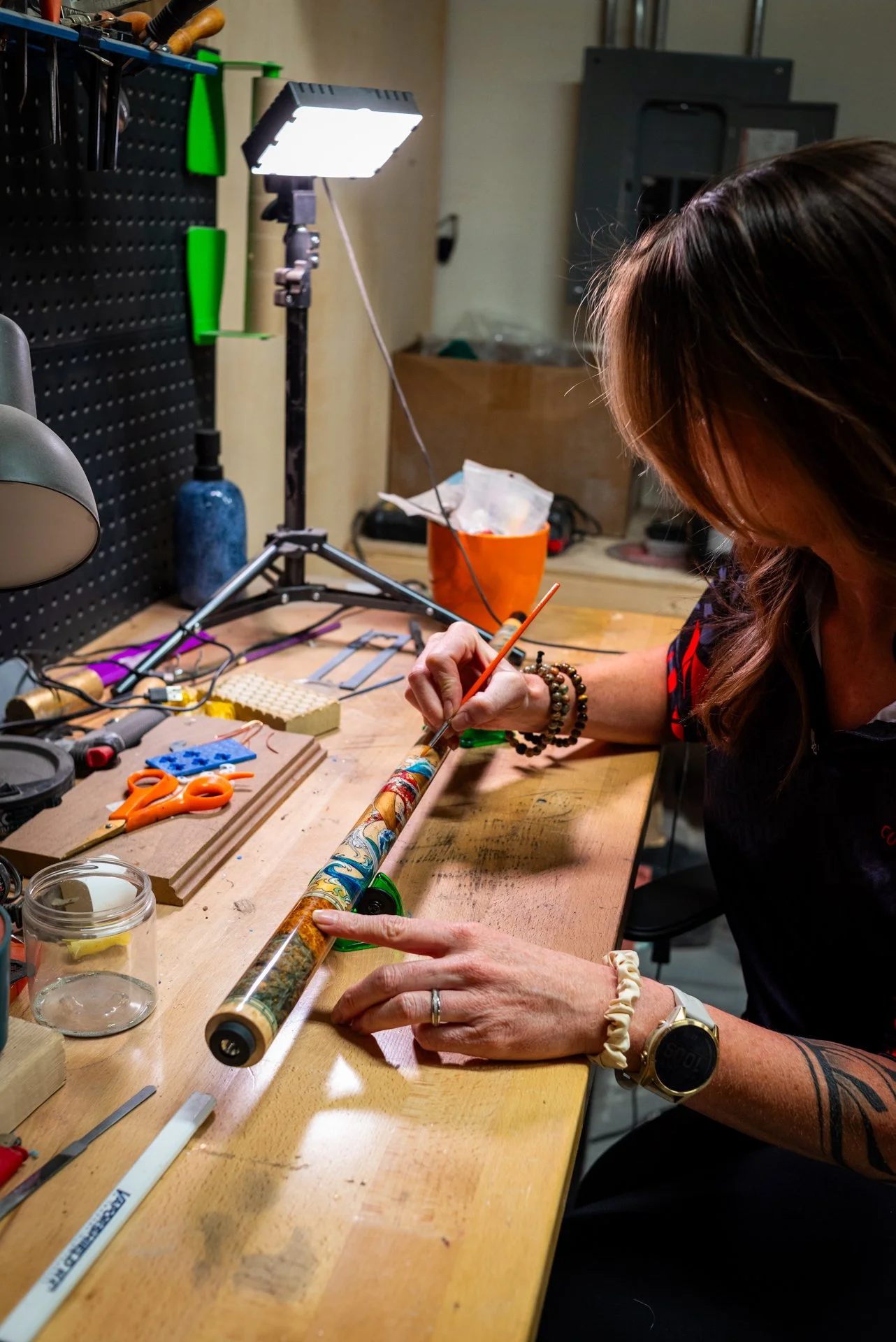 Kaine Cues artist Dawn painting a pool cue with colorful patterns, sitting at a workshop table with her supplies, under a bright light.