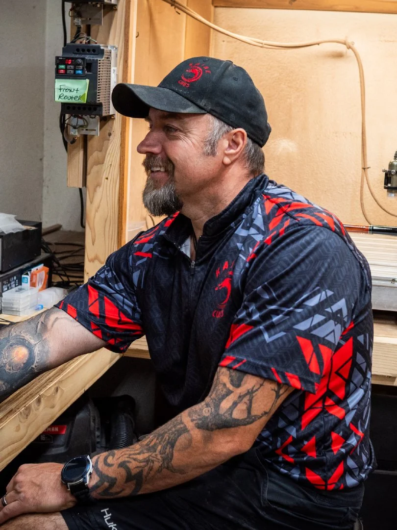 A man with tattoos, a beard, and a black cap sitting at a workbench, smiling, in a workshop or utility room.