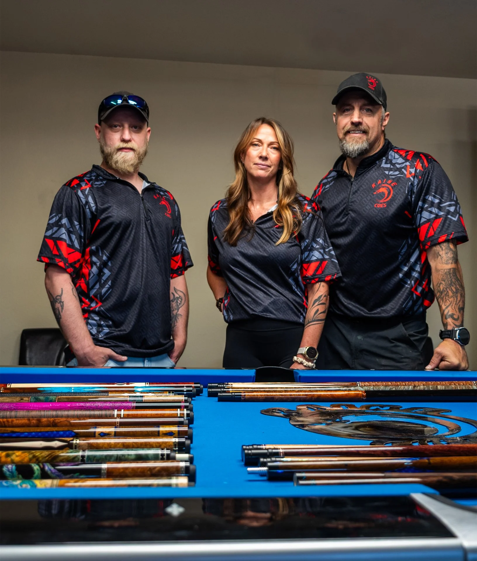 Three people standing behind a pool table with pool cues laid out on it. They are wearing matching black, red, and blue sports jerseys. The man on the left has a beard, tattooed arms, and sunglasses on his cap. The woman in the middle has long wavy hair, and the man on the right has a beard, tattooed arms, and is wearing a cap.