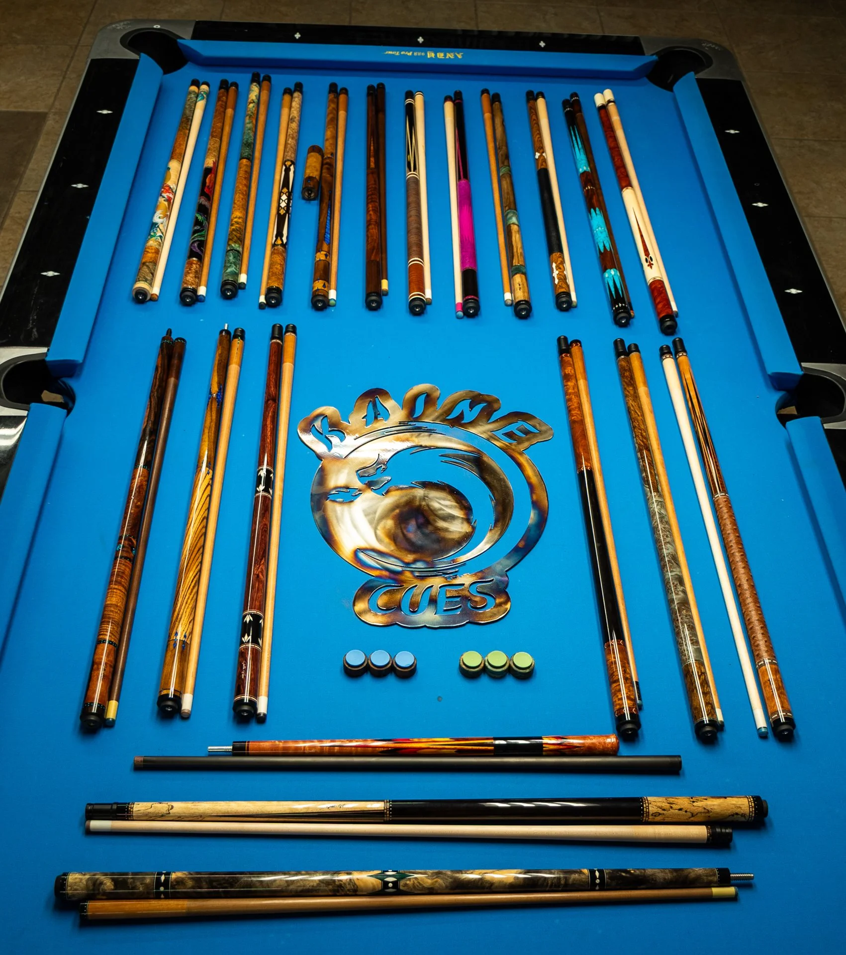 A blue pool table featuring a collection of cue sticks with various wood finishes, patterns, and colors arranged on its surface, along with several chalks at the bottom and a metal decorative piece, the word 'BARGE' in the center, at a game or collection display.