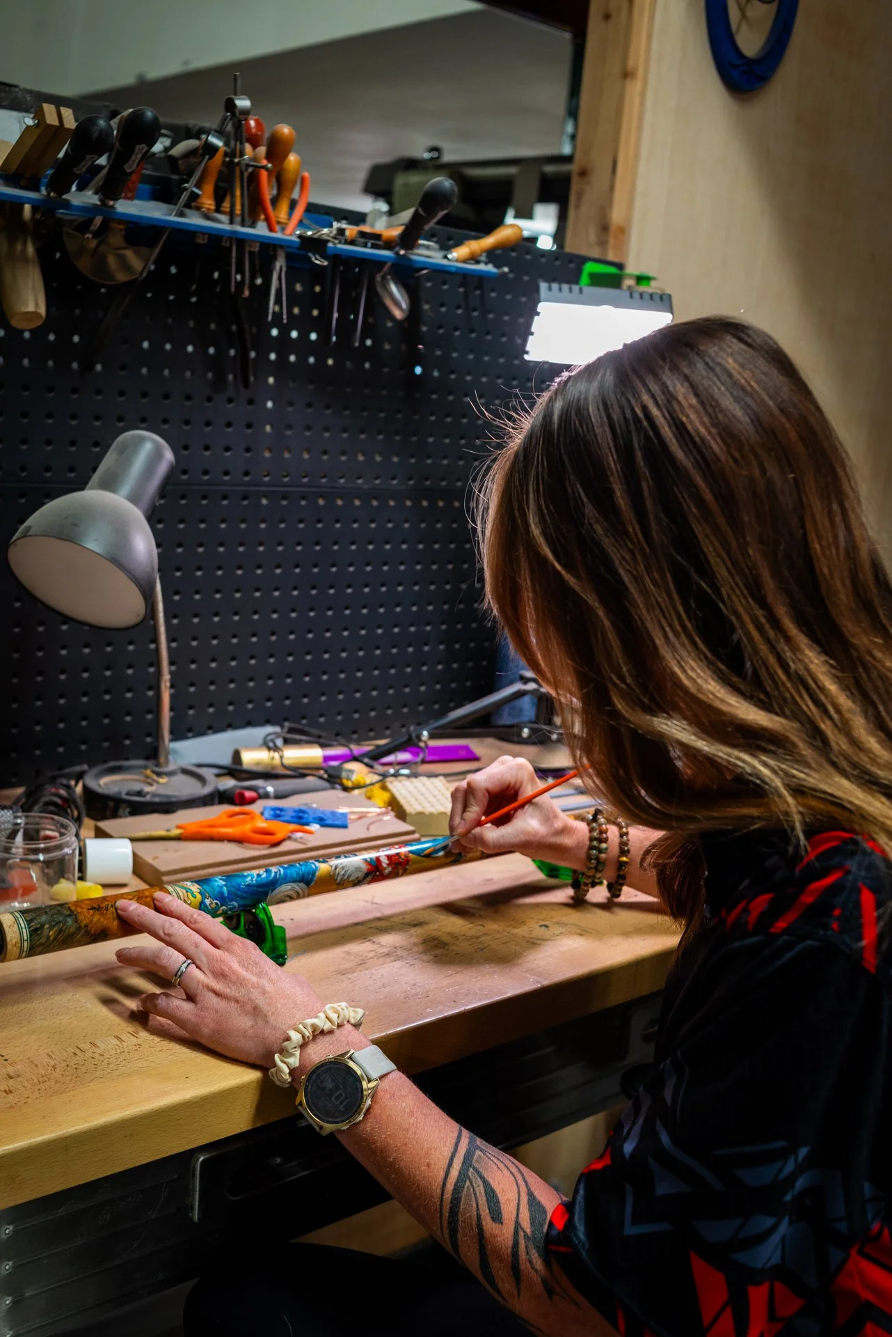 A woman painting a pool cue at a workbench, surrounded by tools and supplies in a workshop.