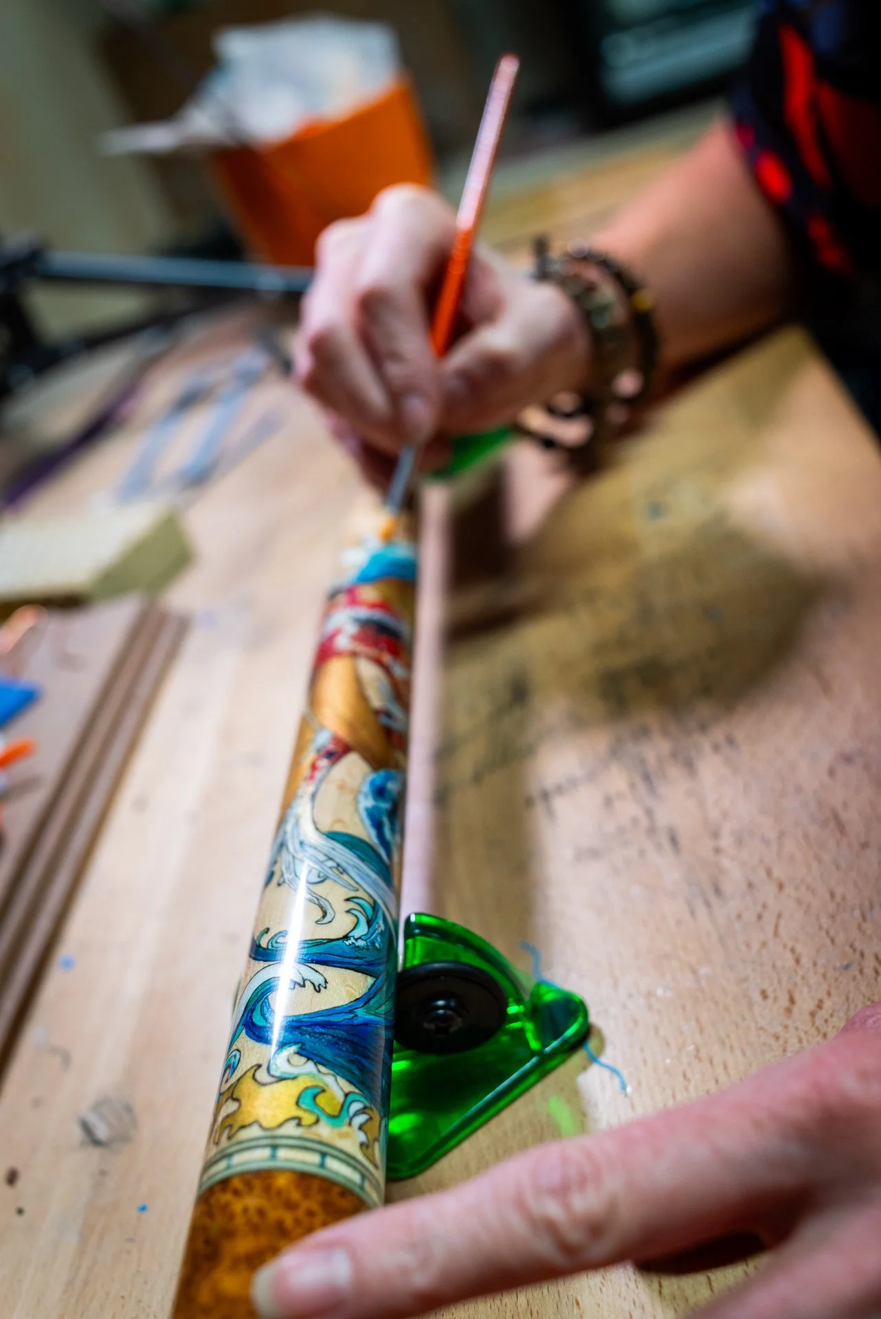 Close-up of a hand painting a colorful design on a pool cue on a workbench.