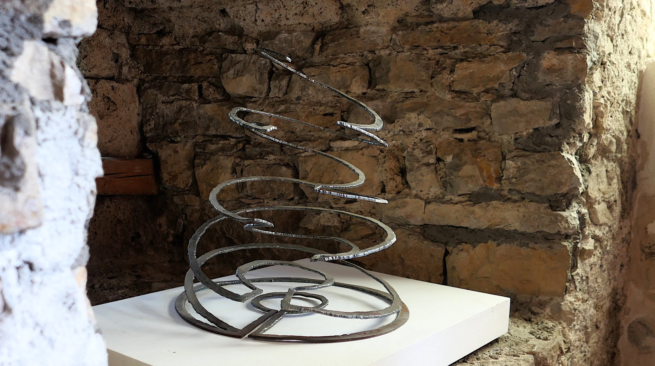Metallskulptur in Spiralform vor einer Steinwand in einem rustikalen Raum, präsentiert auf einer weißen Plattform.
