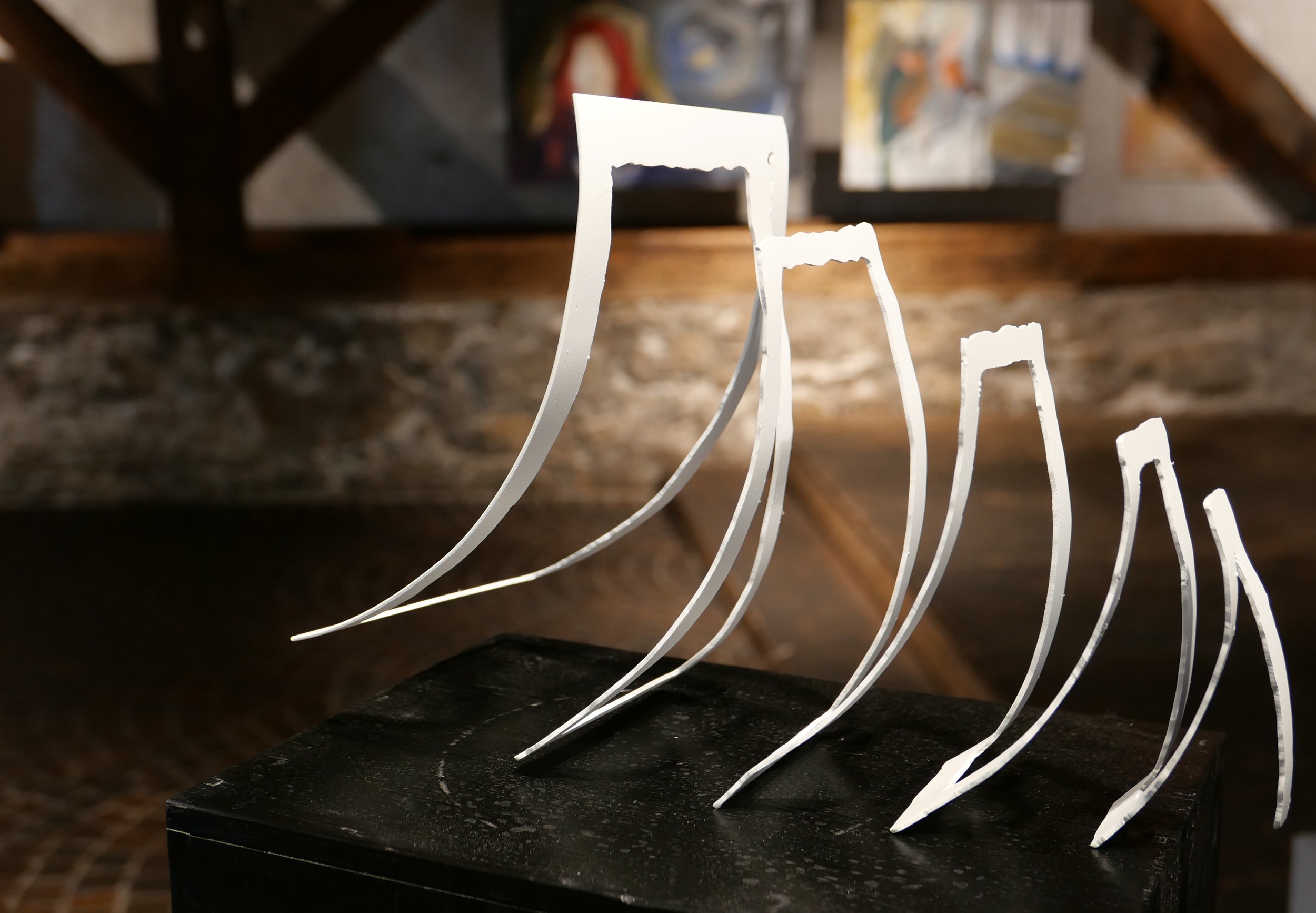 Moderne Skulptur aus gebogenen, weißen Metallstreifen, die parallel angeordnet sind. Im Hintergrund sind unscharfe Kunstwerke an einer Steinwand sichtbar.