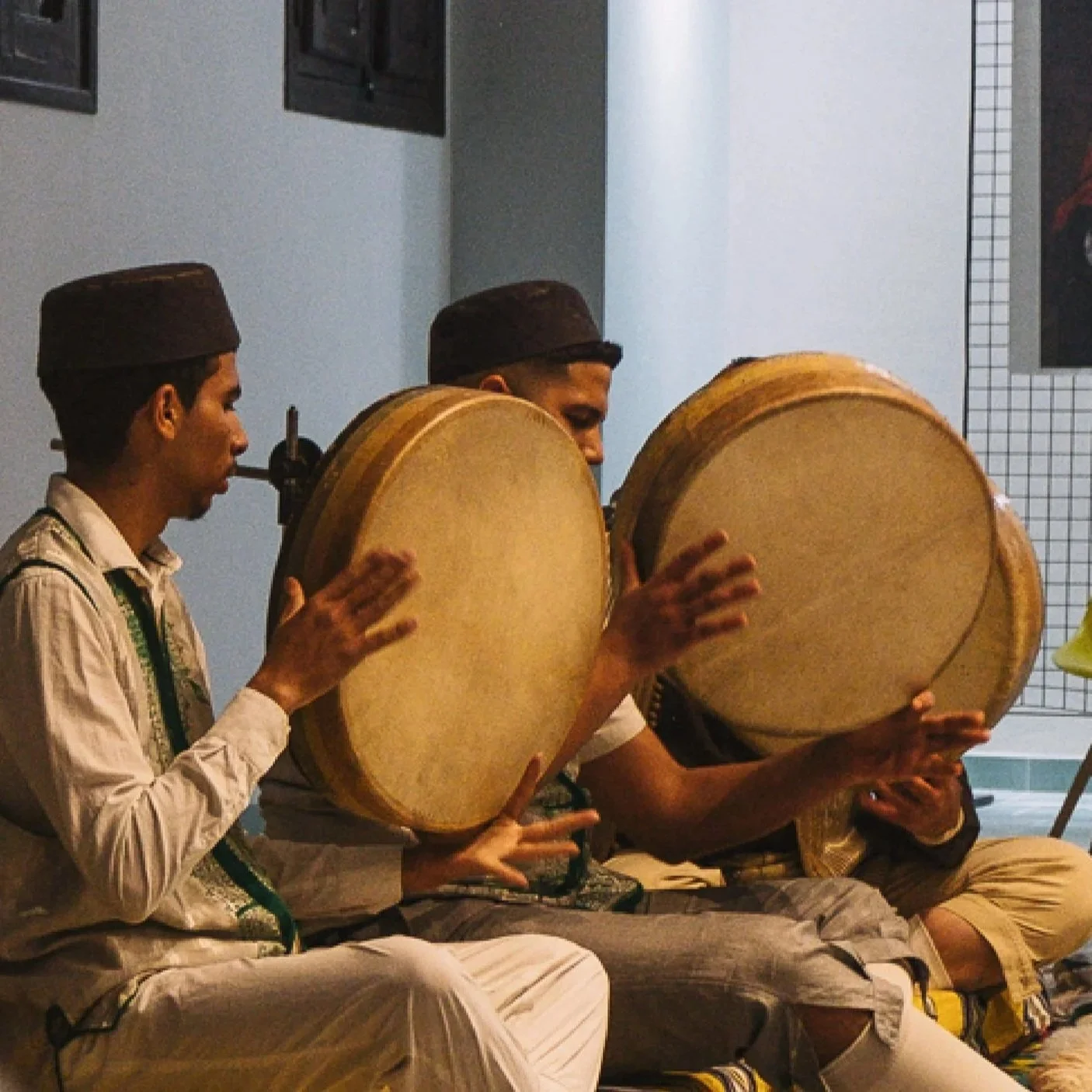 Three young men playing traditional drums while sitting on the floor, dressed in traditional attire, in an indoor setting.