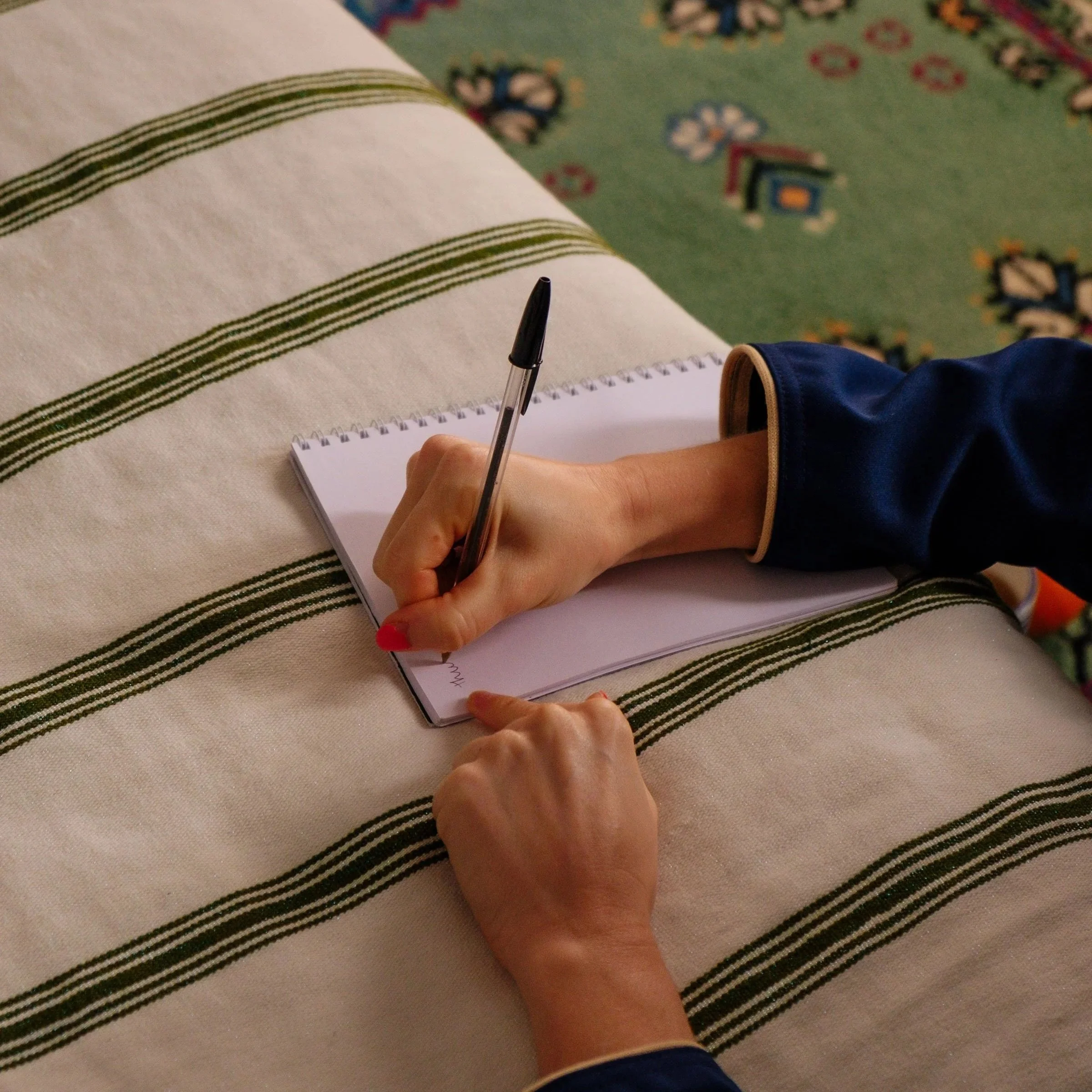 A person with red-painted nails writes on a spiral notepad with a pen, resting on a striped tablecloth. The person is wearing a dark-colored long-sleeve shirt, and the background features a patterned carpet.