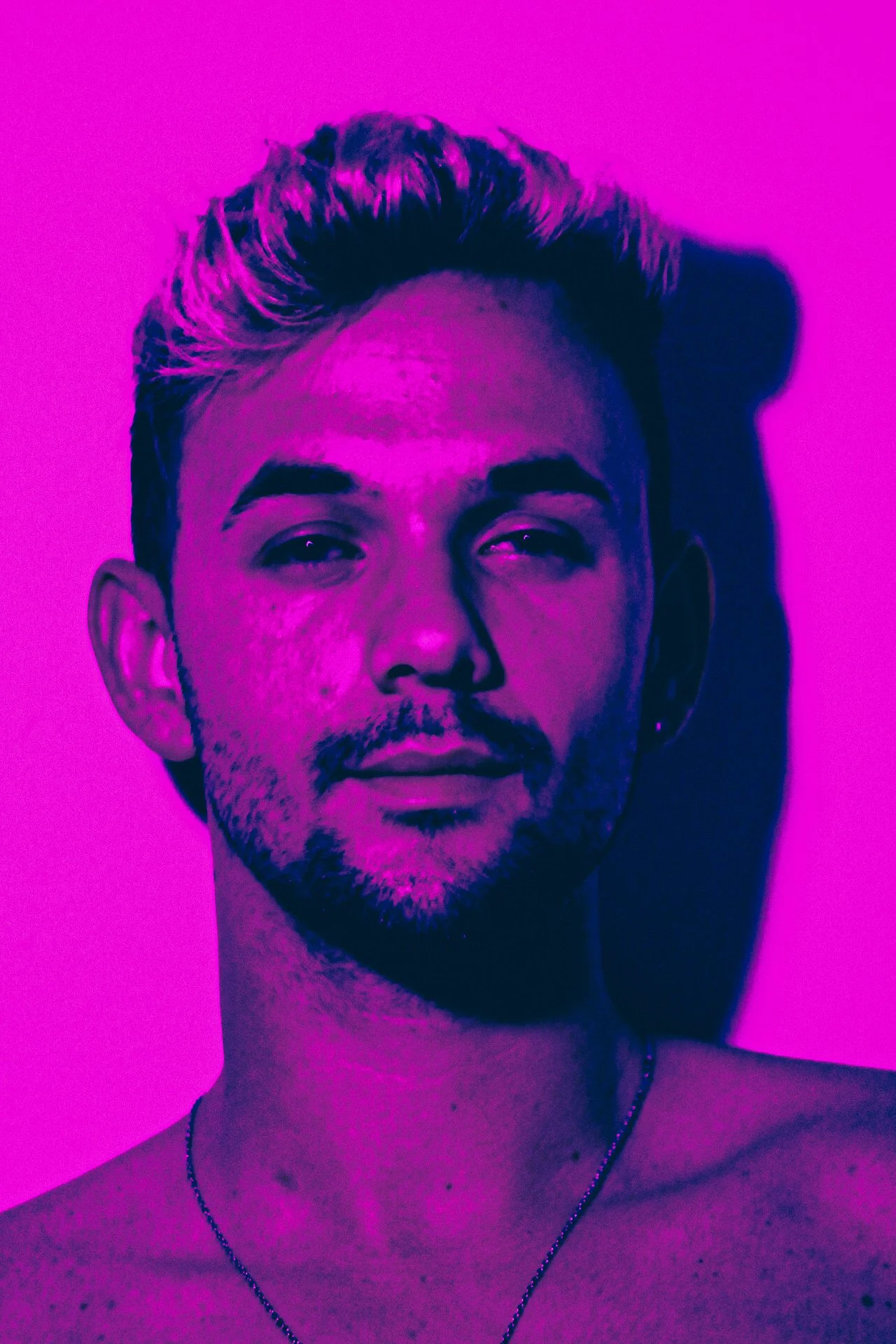 A young man with short hair and facial hair is illuminated by purple lighting, with a serious expression, against a pink background.