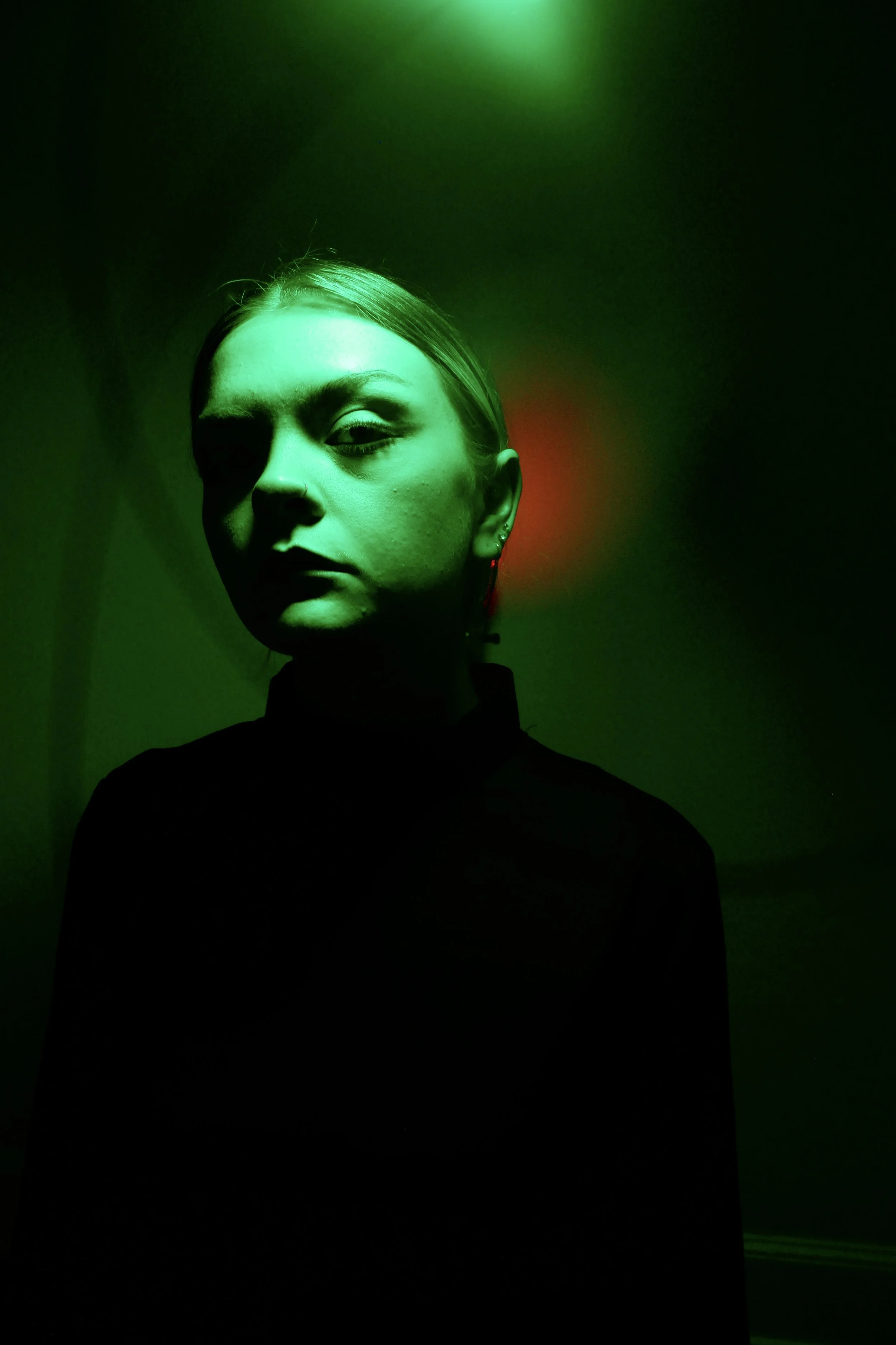 Portrait of a woman with her face illuminated by green light, wearing a dark top. The background is dark with a faint red light behind her ear.