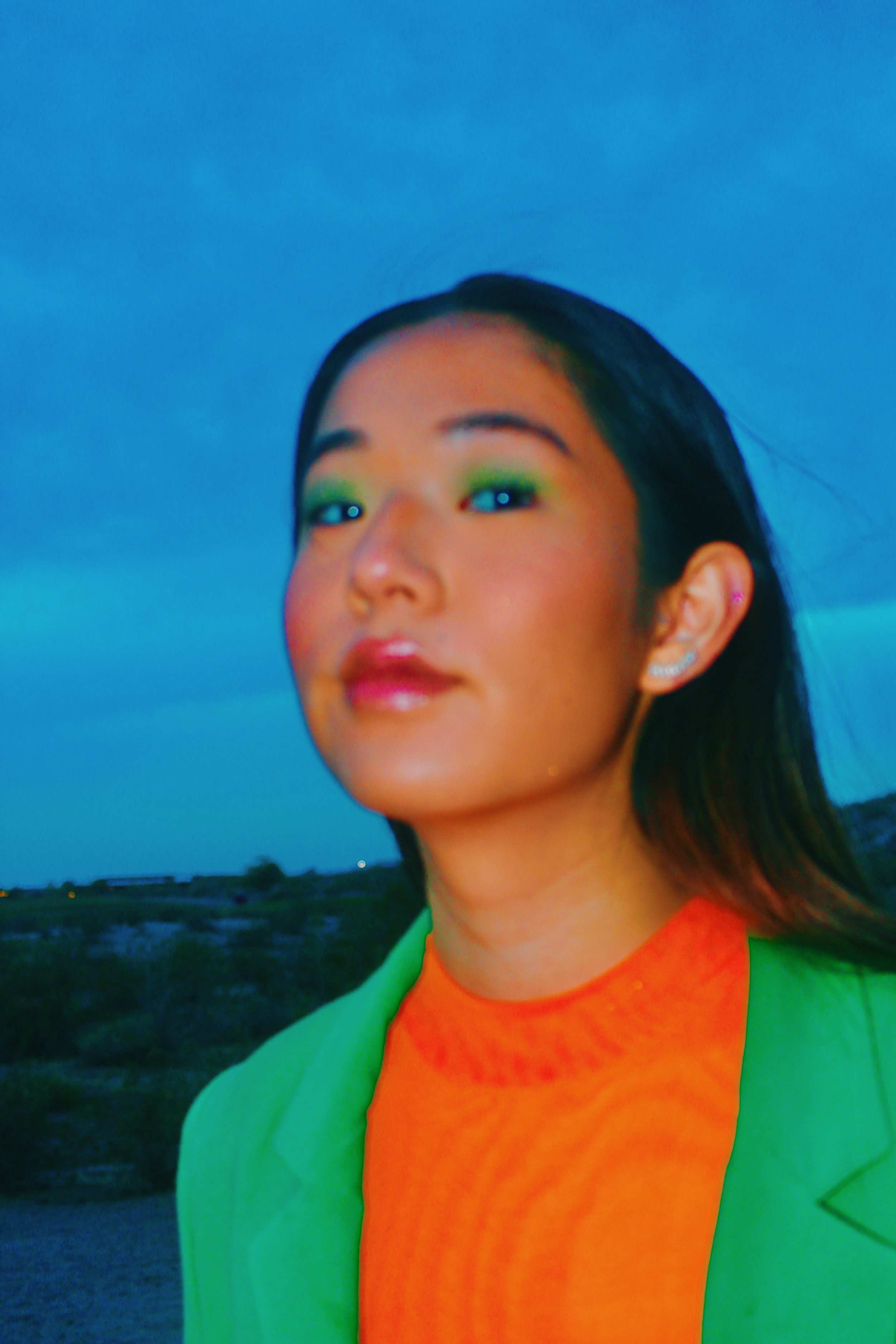 A young woman with makeup, wearing an orange shirt and a green jacket, standing outdoors during twilight with a landscape and cloudy sky in the background.