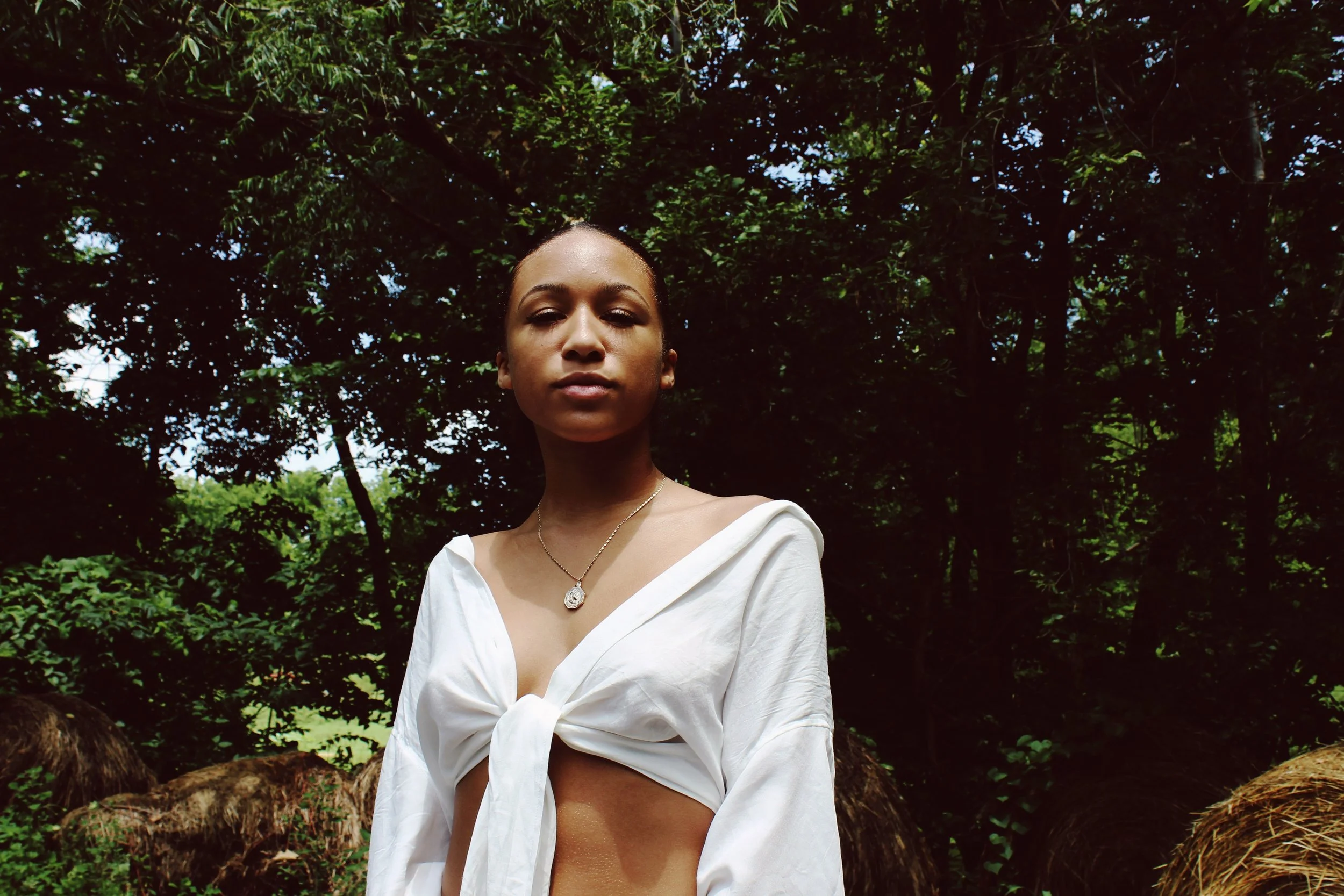 A young woman with dark hair, wearing a white top tied in the front, standing outdoors in a lush green forest with tall trees and hay bales in the background.