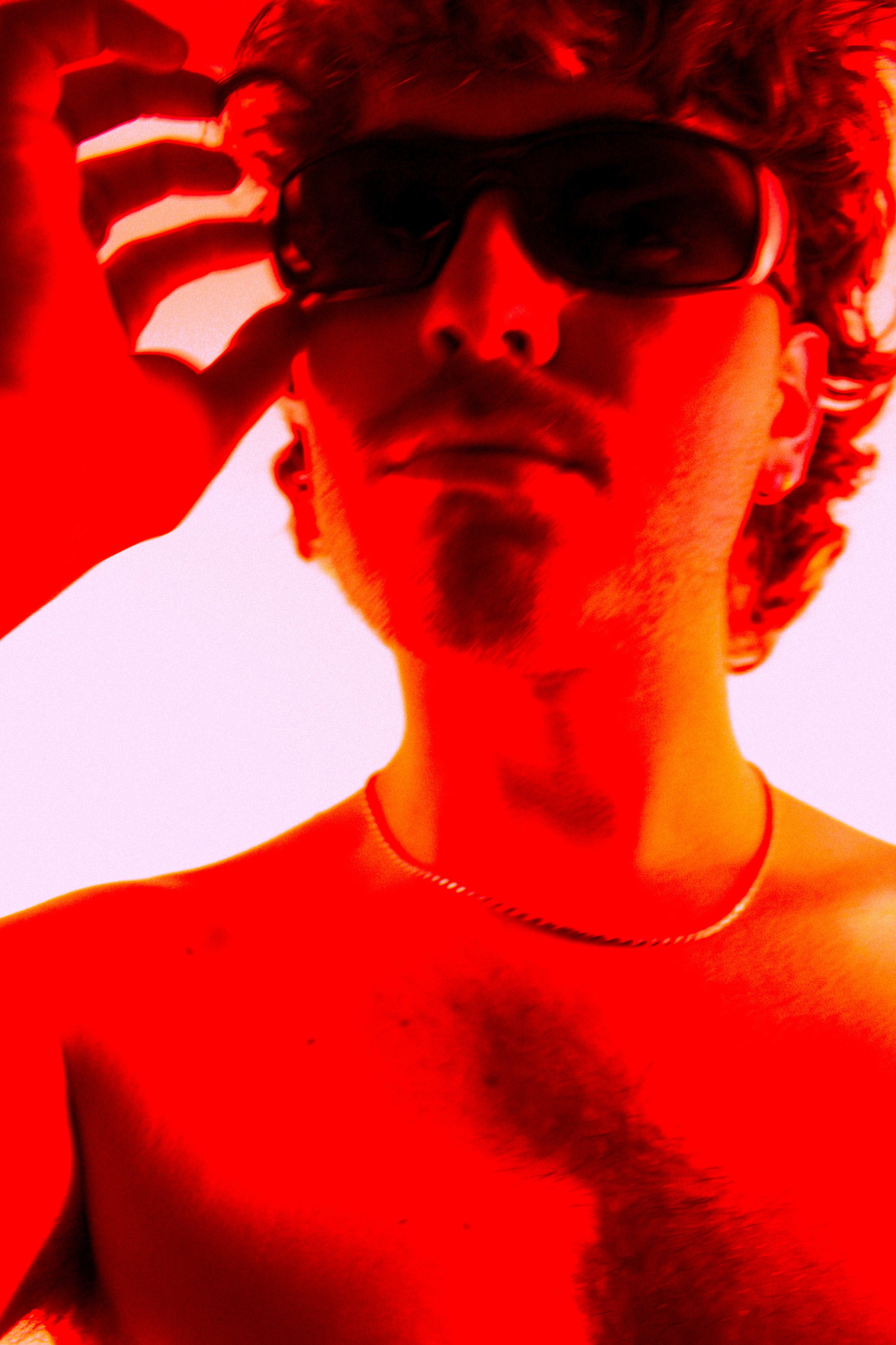 A shirtless young man with a goatee and mustache, wearing sunglasses and a thin chain necklace, posing against a red-toned background.