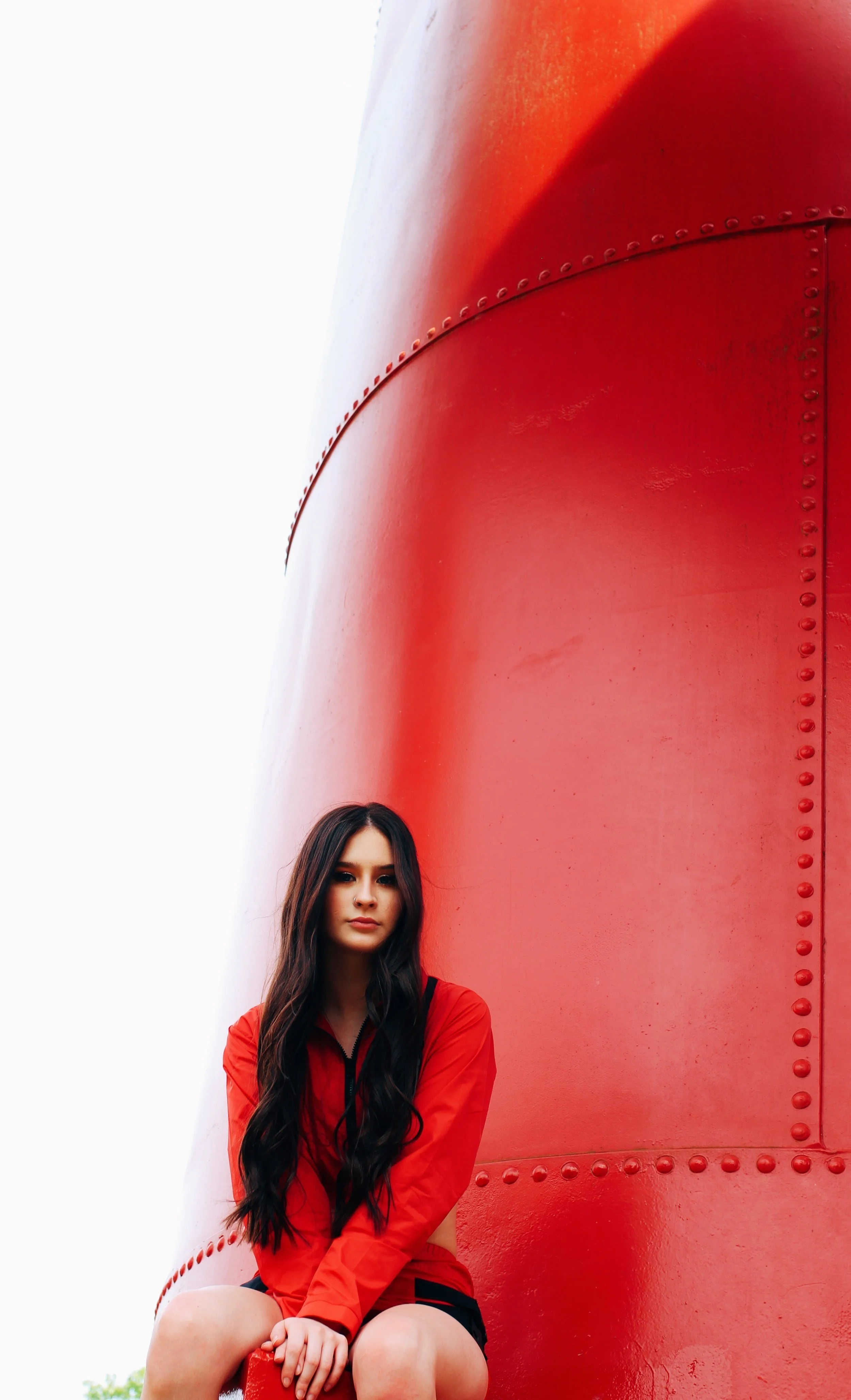 Young woman with long dark hair in a red jacket sitting against a red metal structure with rivets.