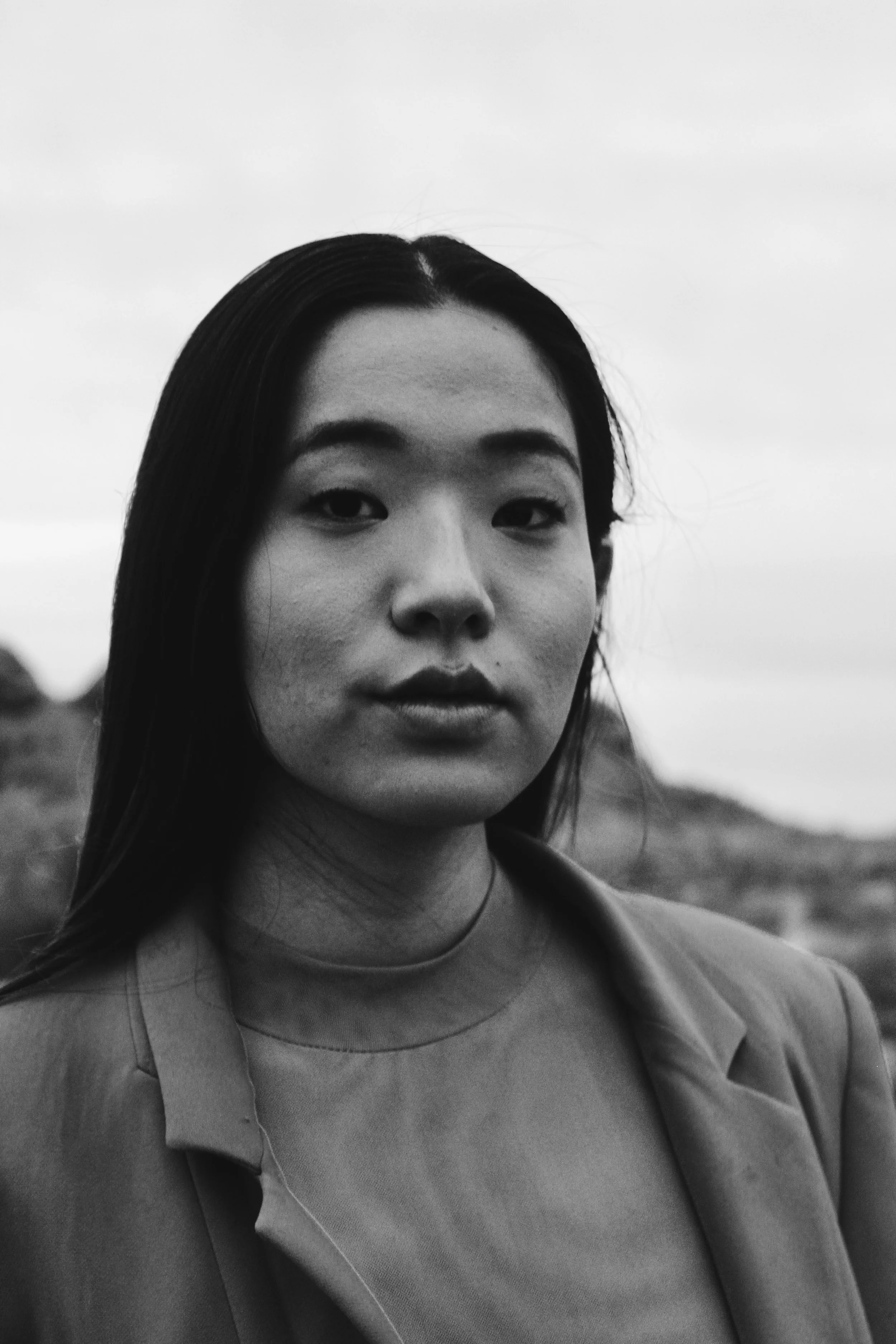 Black and white portrait of a woman with long dark hair, wearing a blazer and round-neck top, outdoors with a blurred landscape background.