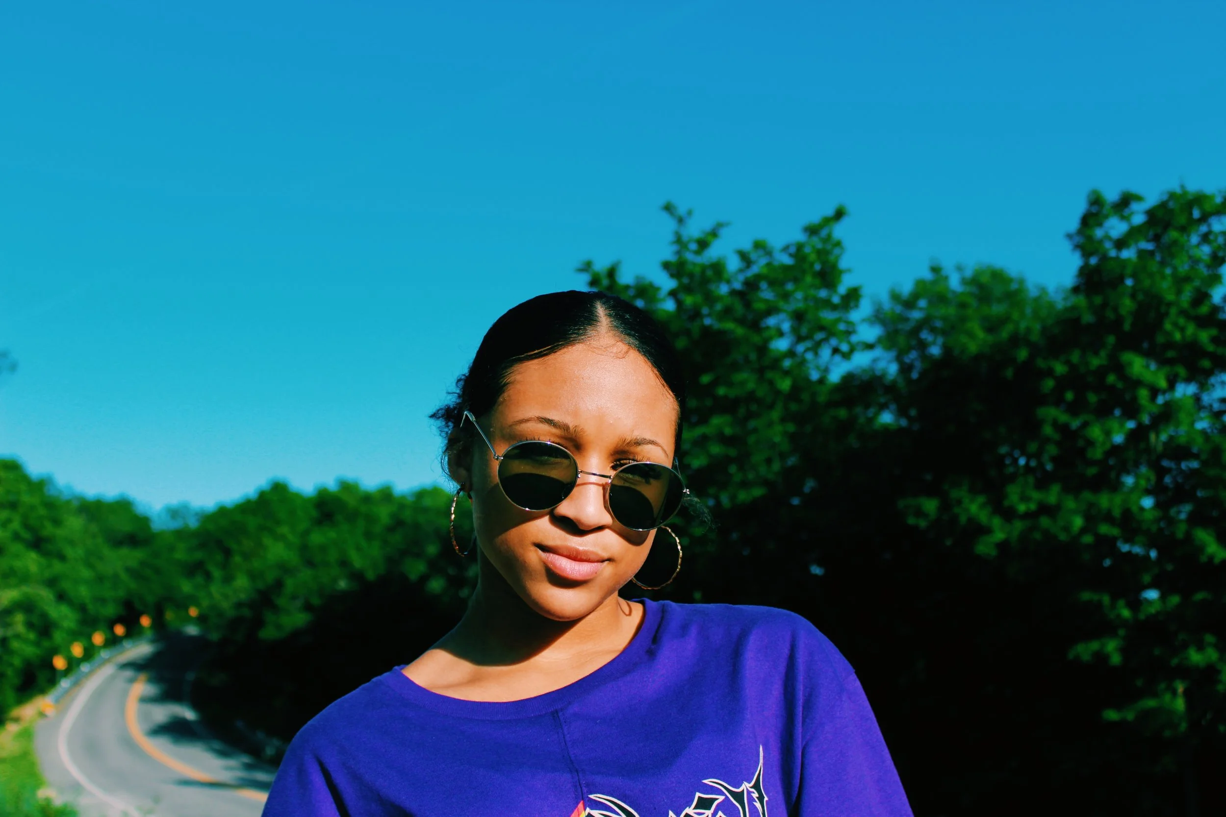 A woman with dark hair wearing round sunglasses and a blue shirt standing outdoors next to a curving road with green trees and a clear blue sky in the background.