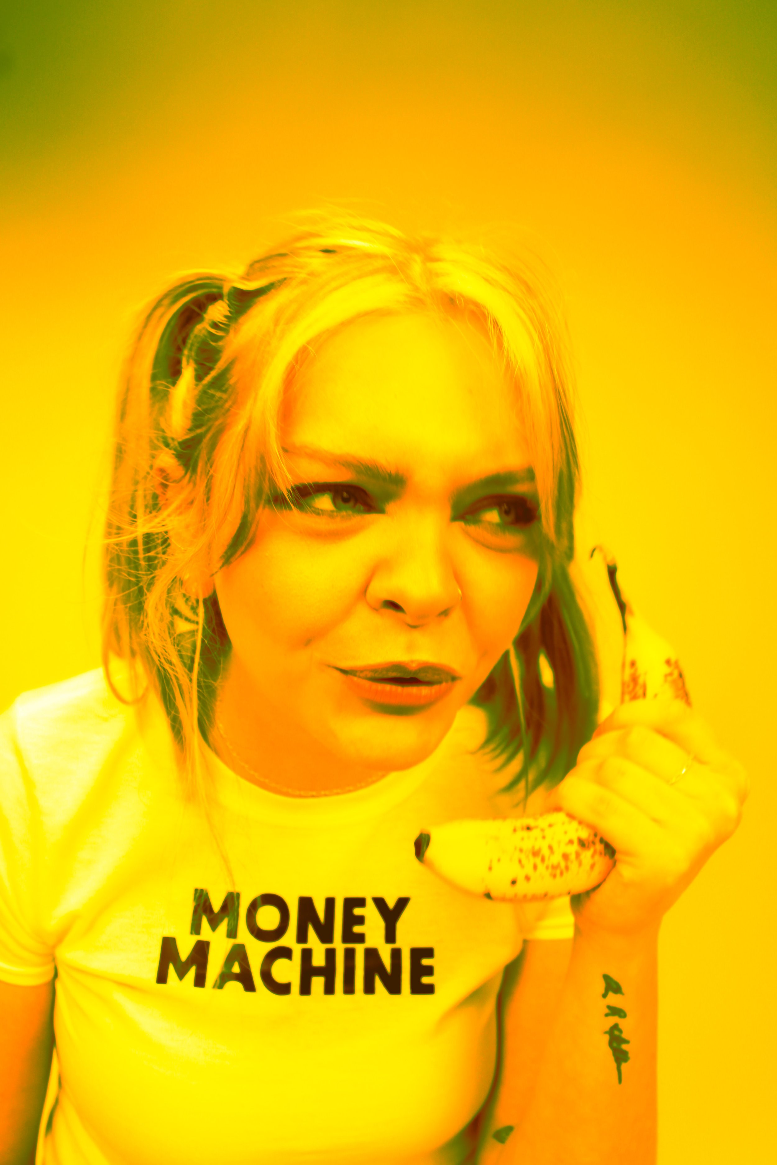 A woman with blonde hair holding a banana to her ear as if it were a phone, wearing a yellow T-shirt with black text 'MONEY MACHINE', against a yellow background.