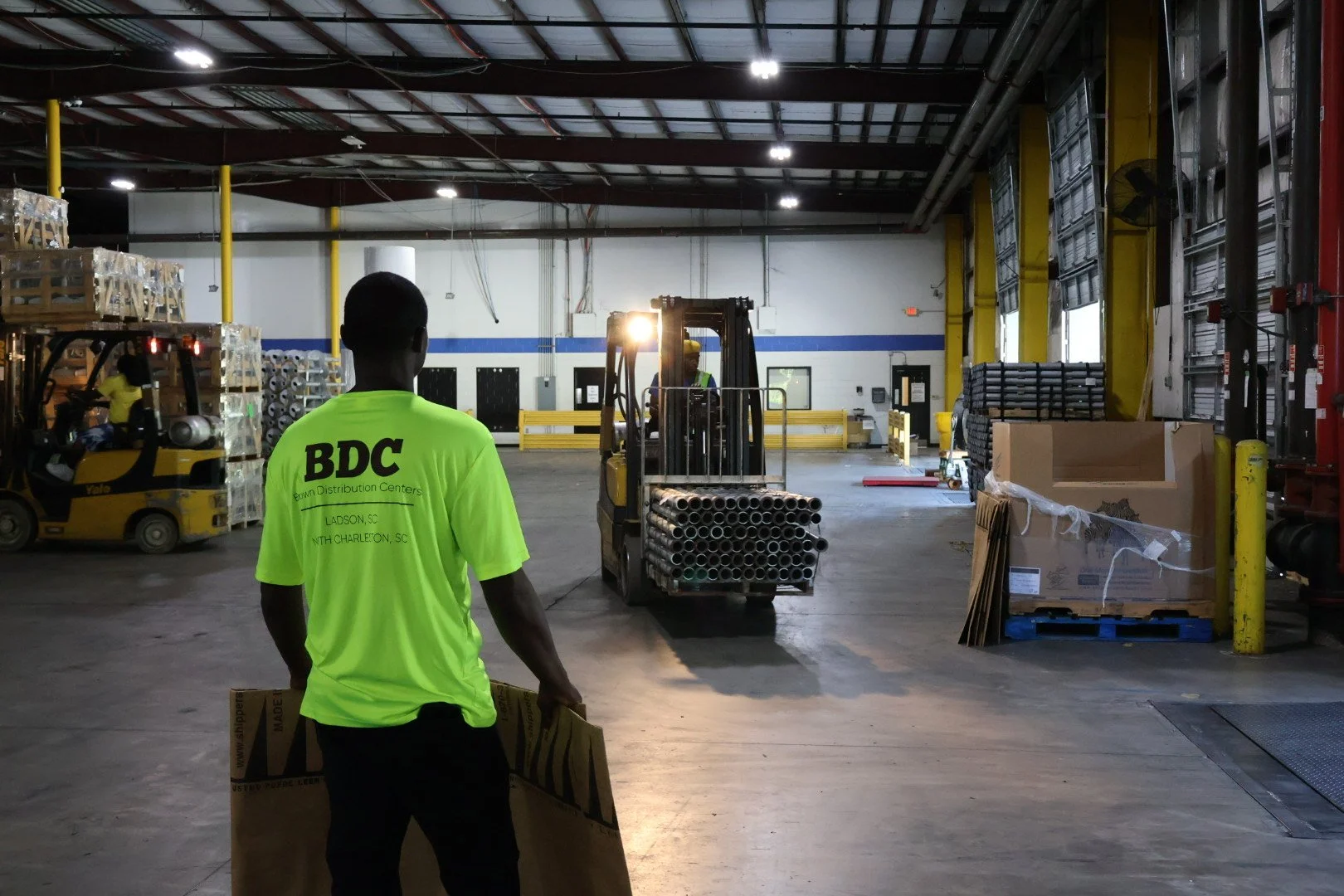Worker in a bright yellow shirt with 'BDC' on the back stands inside a warehouse, holding a cardboard box. A forklift with pipes is in motion in the background, with other warehouse equipment and shelves around.